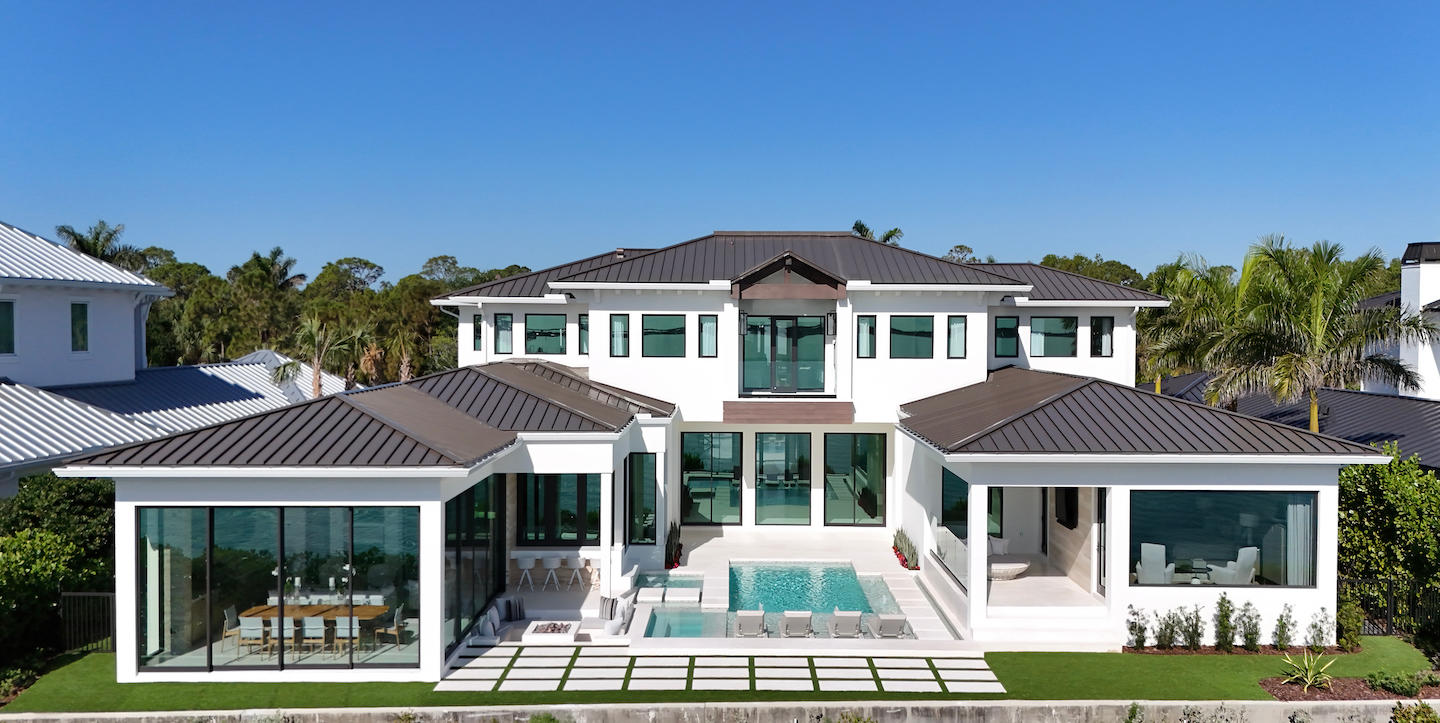 Drone view of a bright white coastal estate capped with sleek dark metal roofs, its symmetrical single-story wings flanking a taller central volume. Floor-to-ceiling glass doors open to a courtyard with a lap-style pool, turf-inlaid paver paths, and rows of sun loungers. Inside, a glass-walled dining room and lounge are visible, all set against palms and a flawless blue sky.