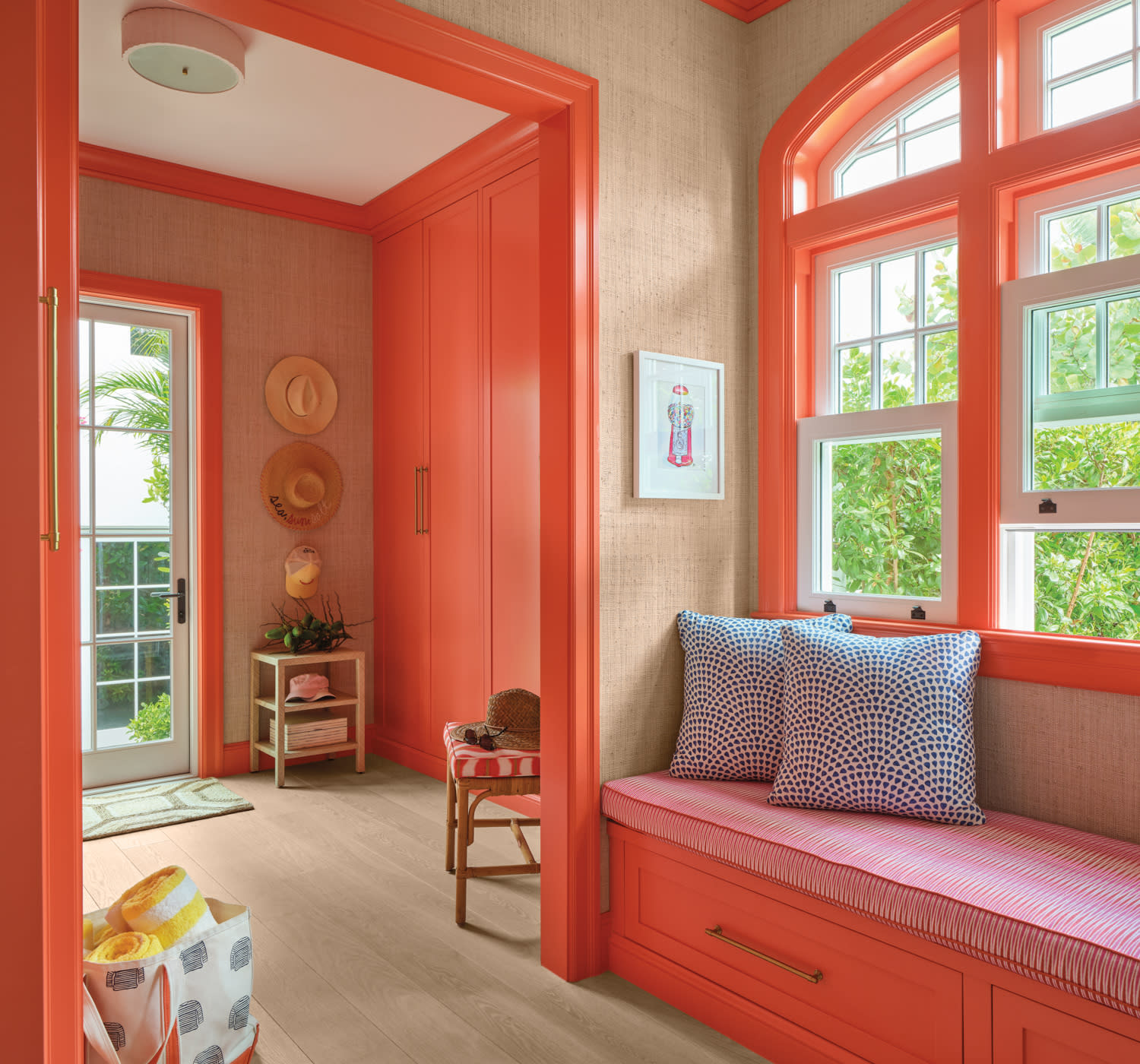 Bright, coral-colored entryway with a large bench, patterned cushions, and decorative hats. Natural light streams through a window, creating a warm ambiance.