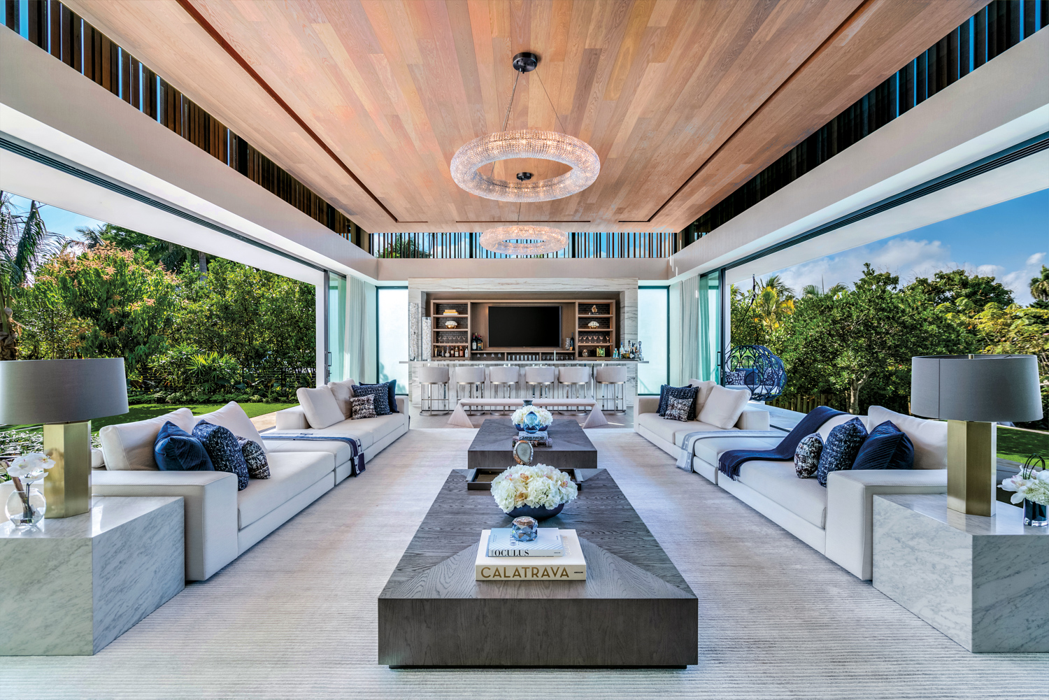 Long, low-profile sofas line an indoor-outdoor space with a bar at one end beneath wood-paneled ceiling.
