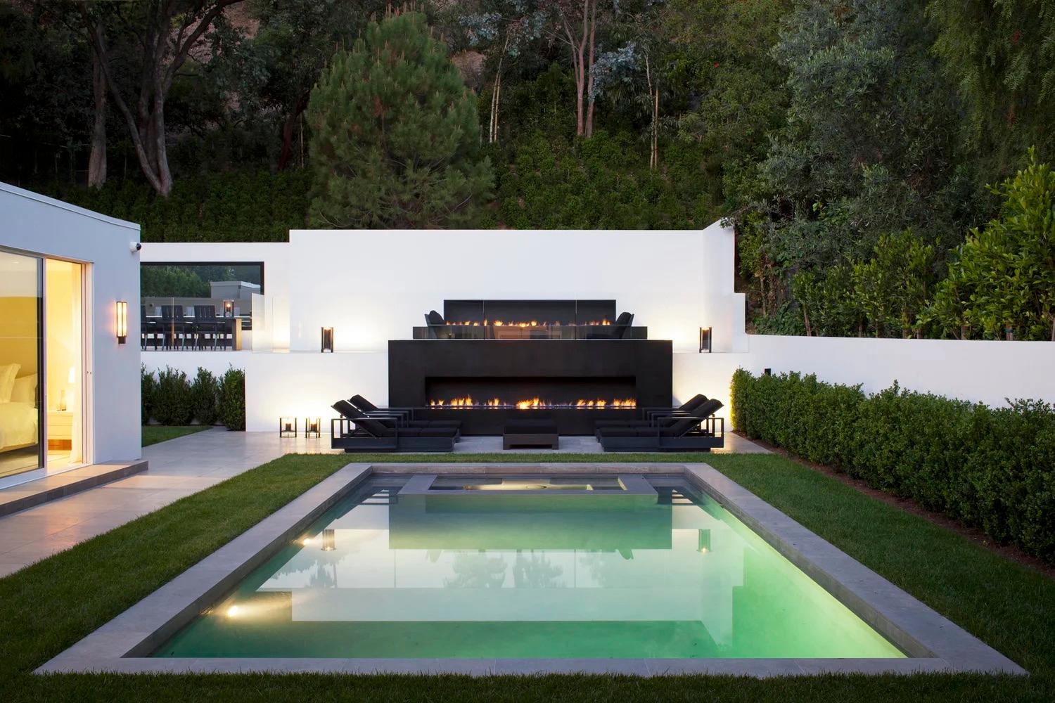 Modern backyard with a sleek rectangular pool, surrounded by manicured grass. Features a stylish outdoor fireplace, lounge chairs, and lush greenery.