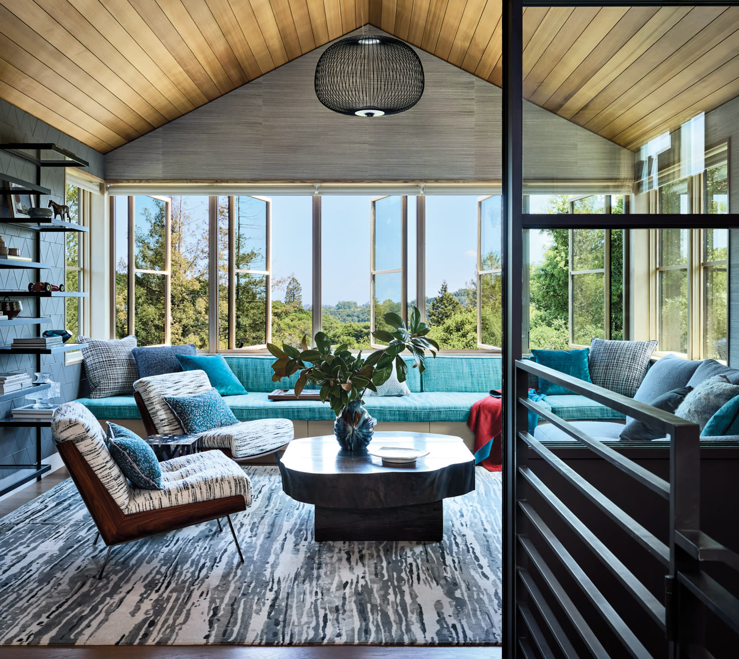 Stylish living room with large windows overlooking greenery. Features teal sectional, patterned chairs, wood coffee table, and modern decor. Inviting and serene ambiance.