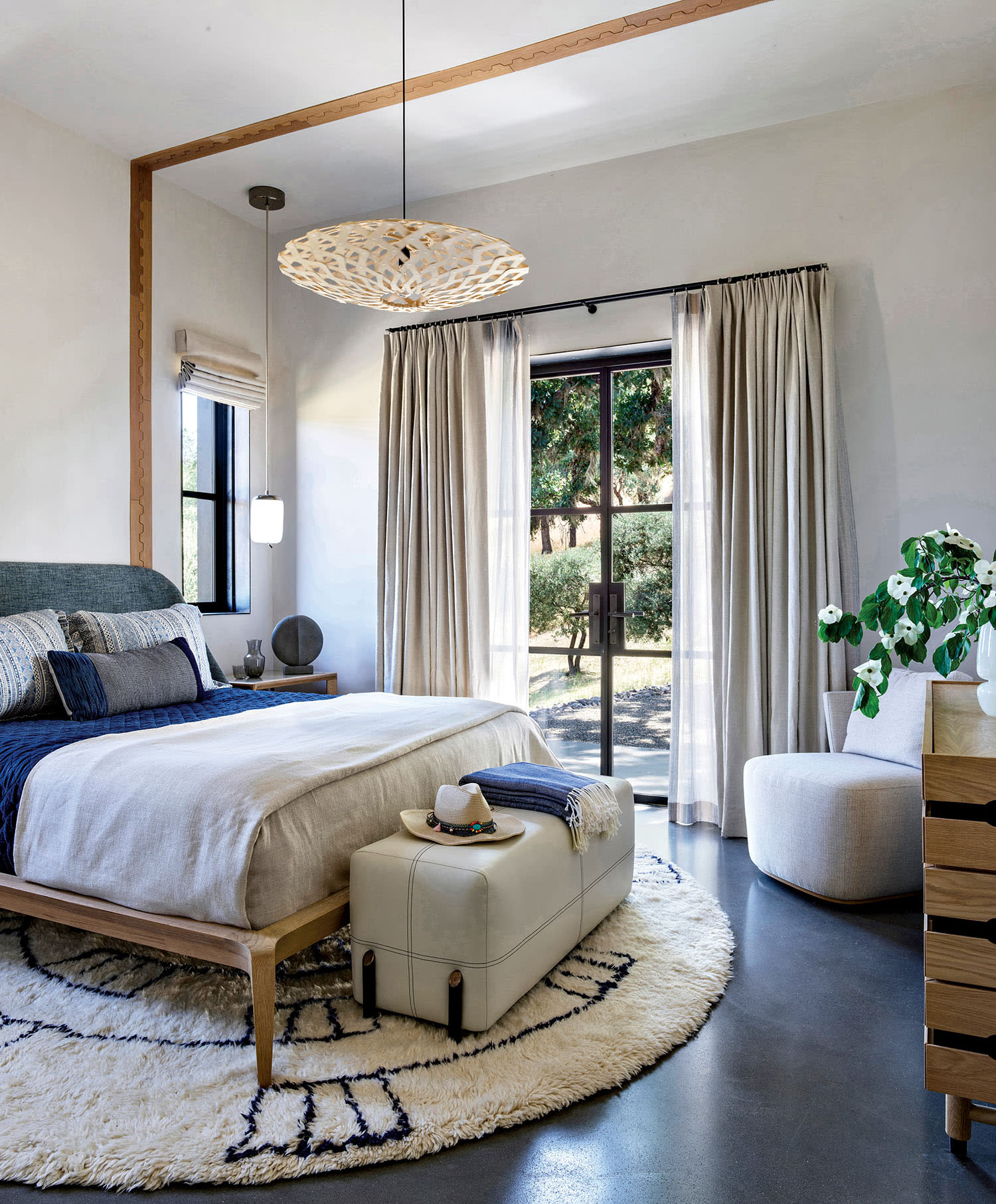 A serene bedroom features a plush bed with blue and beige bedding, a modern pendant light, large window with curtains, round rug, and a leather ottoman.