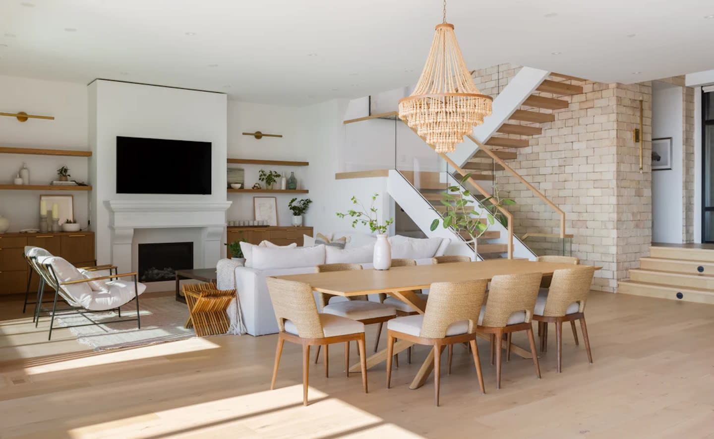 Spacious modern living room with light wood flooring, featuring a large dining table, woven chandelier, white sofa, staircase, and TV above a fireplace.