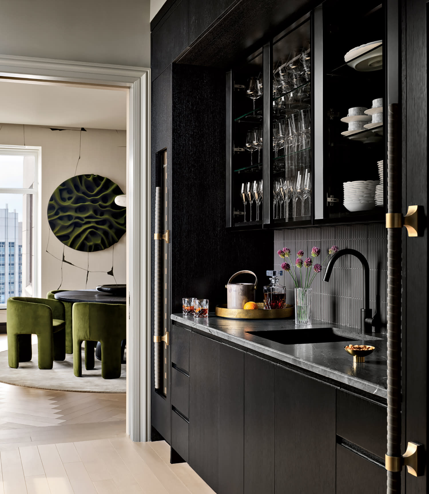 Sophisticated modern kitchen with dark cabinets and glassware, adjacent to a dining area with green chairs and a round wall art. Elegant and stylish.