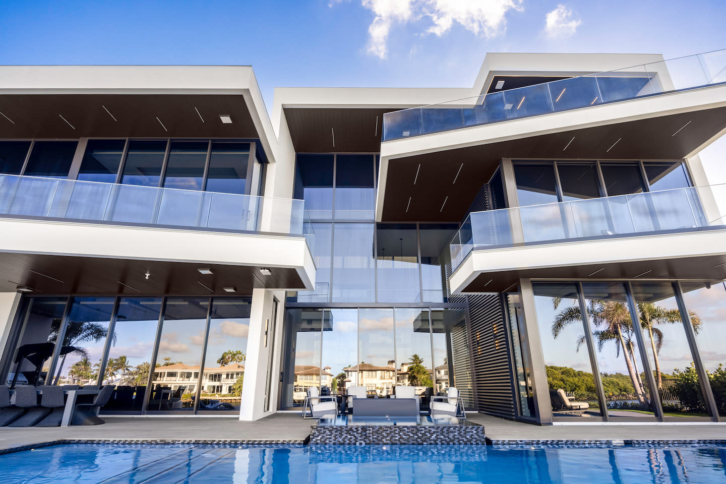 Sleek waterfront mansion captured from pool level, showcasing bold cantilevered balconies with frameless glass railings and soaring double-height walls of reflective glass. Dark wood soffits and slim white frames create sharp geometric lines, while the pool’s blue water mirrors palm trees and neighboring villas beyond an outdoor lounge set.
