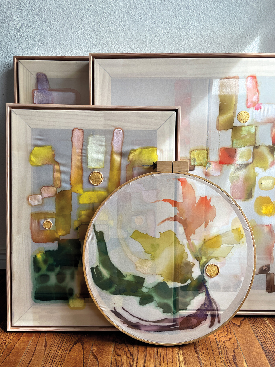 Multiple artworks on silk, including a round-shaped one, by artist Griffin, stacked against a wall