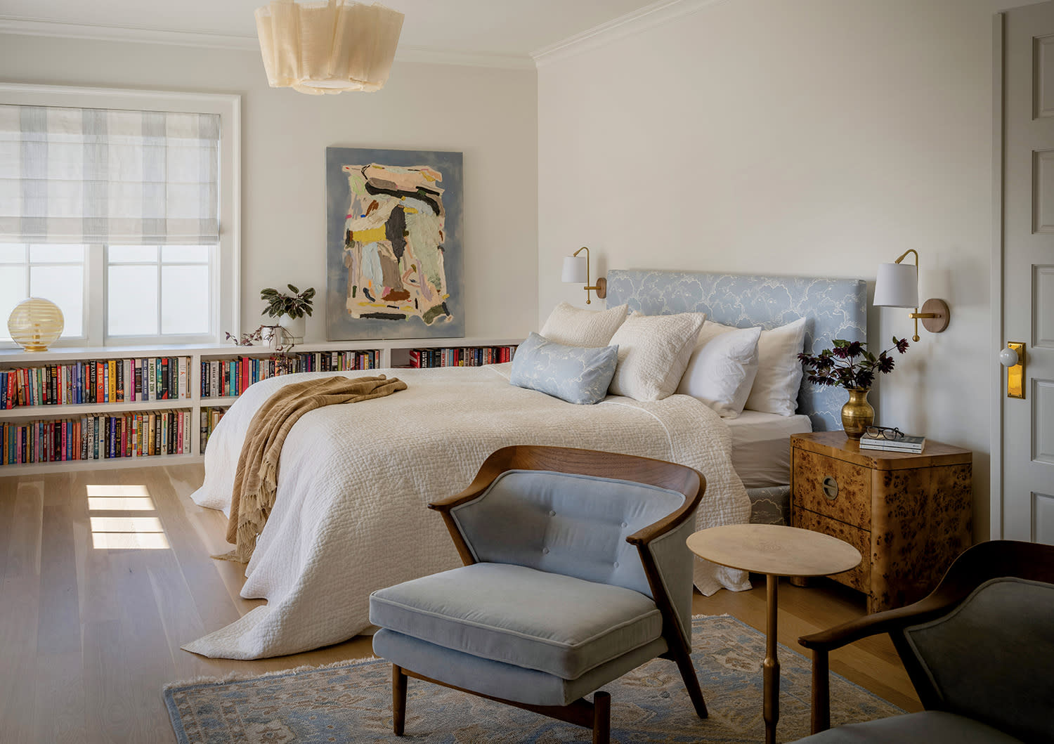 A cozy bedroom with soft lighting features a bed with white linens and blue cushions, a patterned headboard, and bookshelves lining the wall.