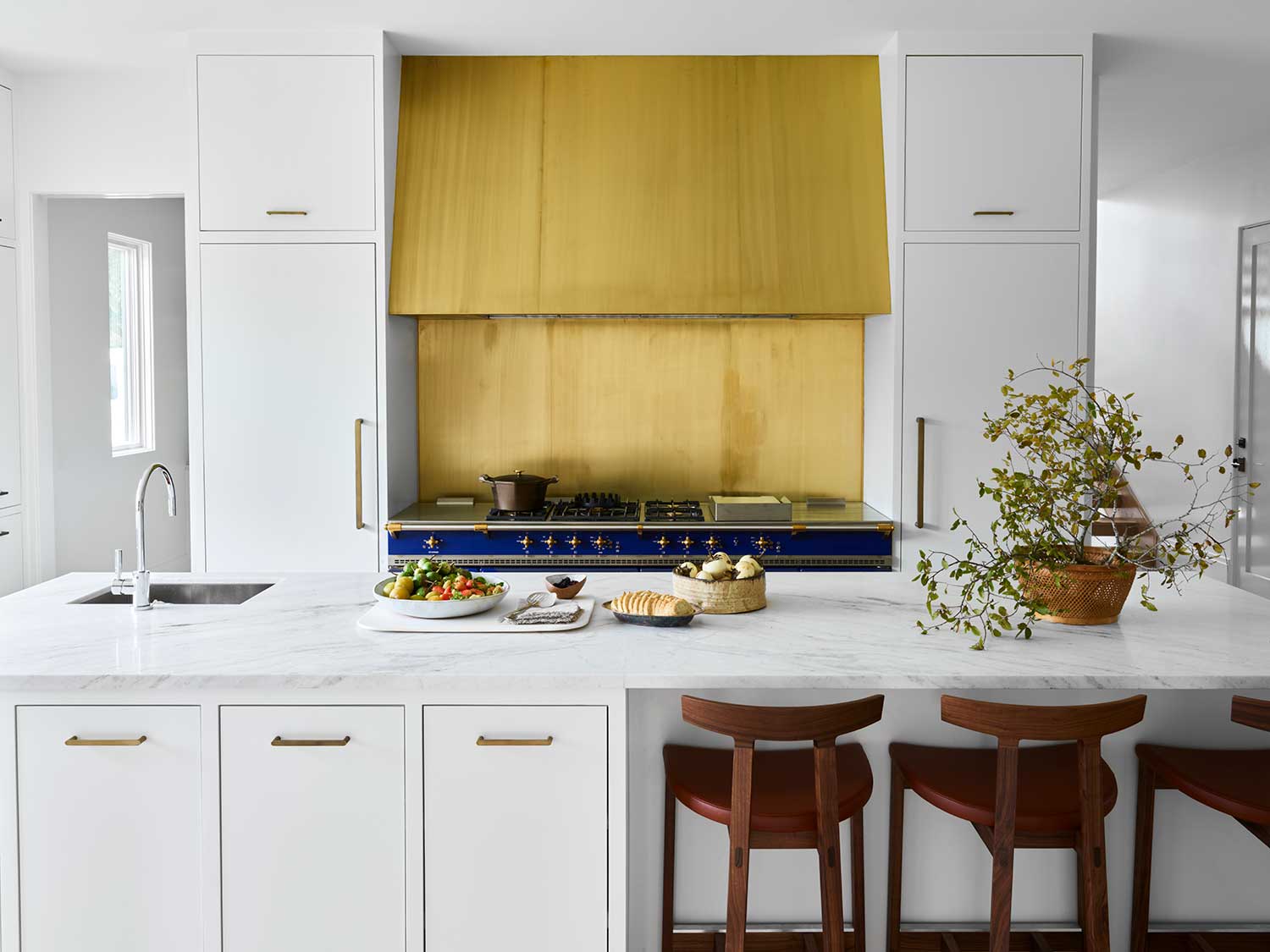 white kitchen with blue range, gold hood, marble island and wood counter stools