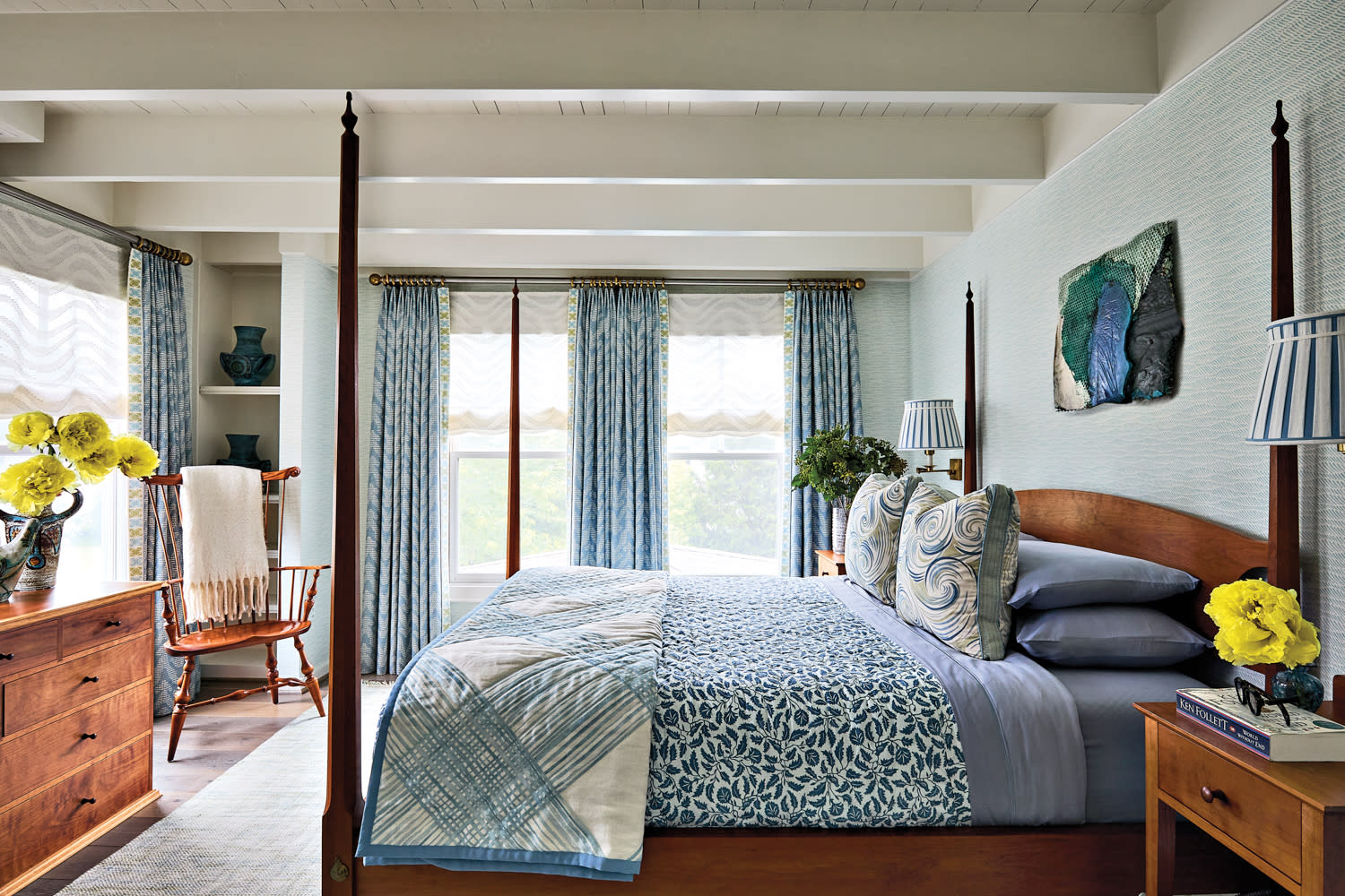 Cozy bedroom with a wooden four-poster bed. Blue floral bedding, matching curtains, and pillows. Bright yellow flowers on nightstands add warmth.