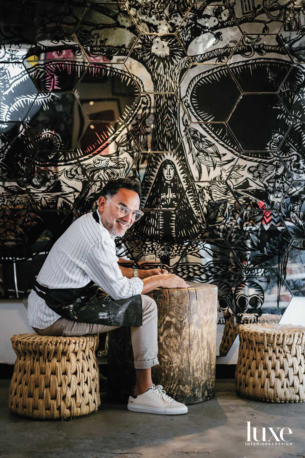 A man sitting on a stool smiling at the camera. Behind him is artwork.