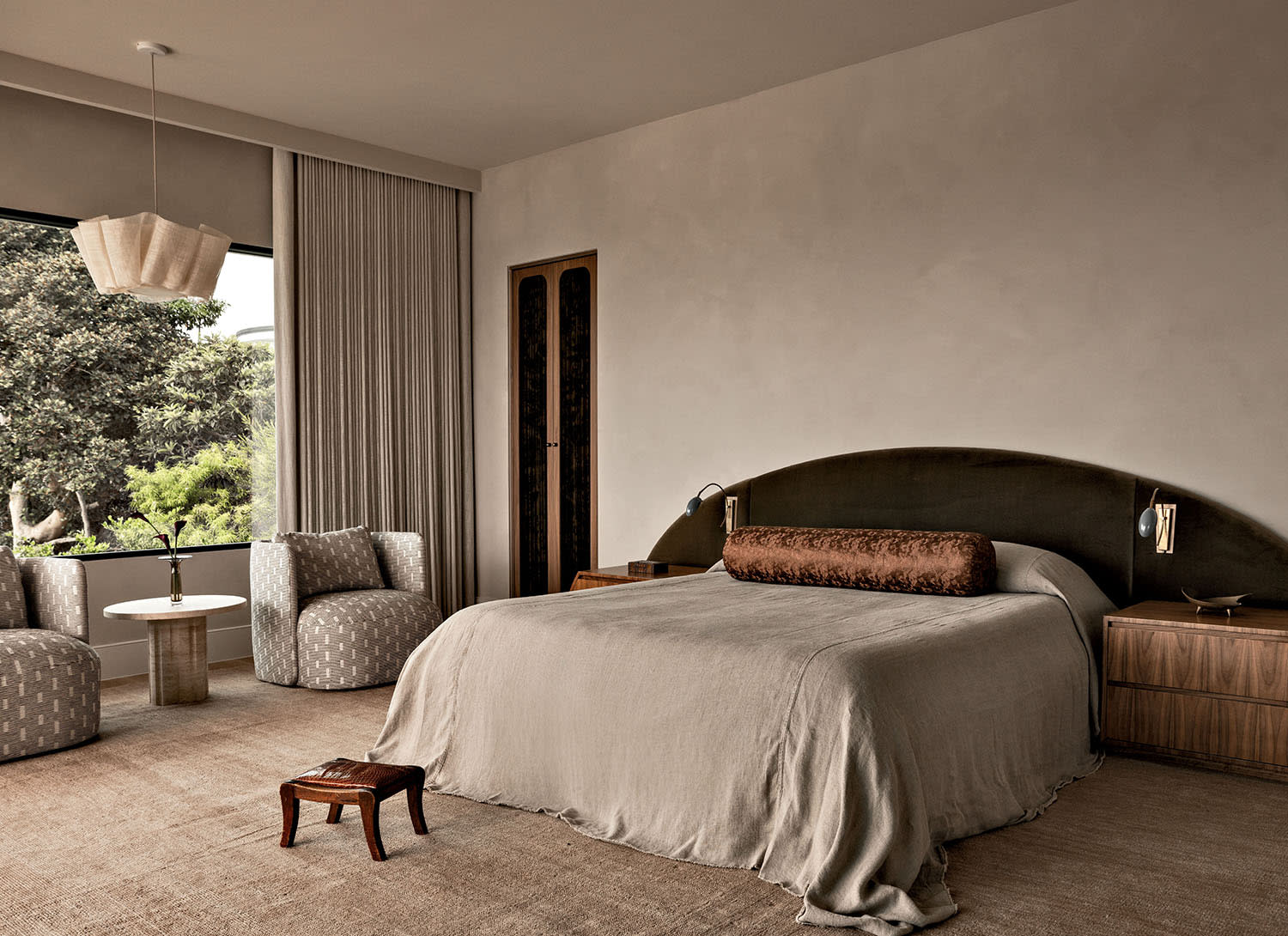 A bed with a brown velvet headboard and wood nightstands, near two armchairs set with a table between them.