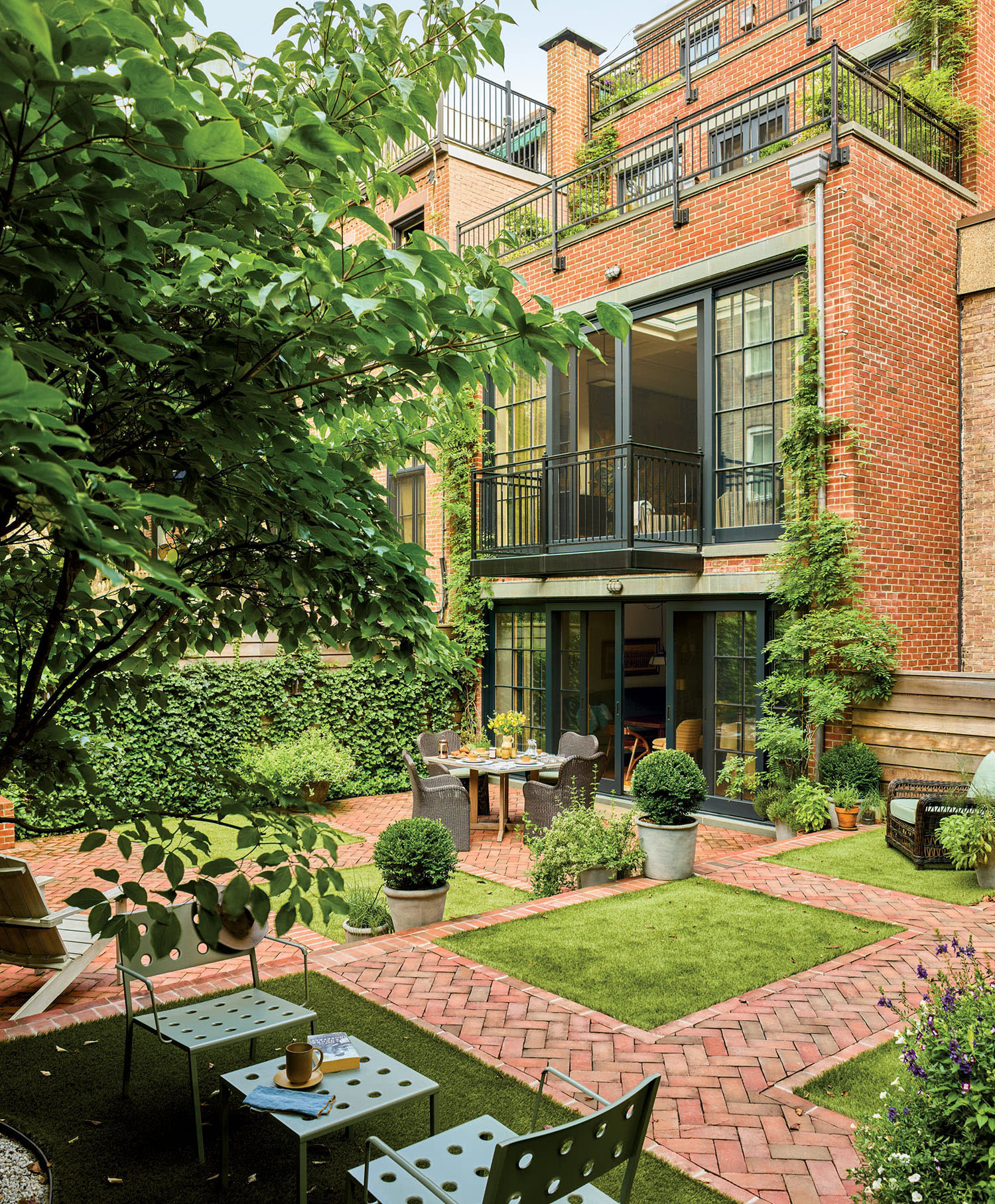 Charming urban garden with lush greenery, red brick paths, and modern seating. A cozy table is set near a brick building covered in ivy. Calm ambiance.