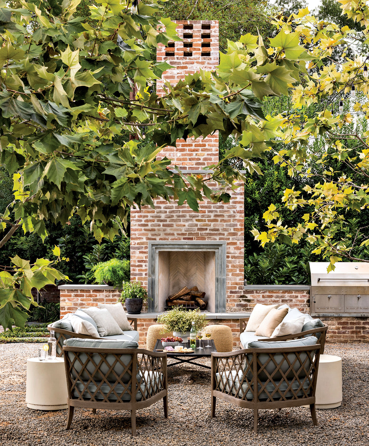 Cozy patio scene with two plush chairs facing a brick outdoor fireplace, surrounded by leafy trees. Casual, inviting atmosphere with a grill nearby.