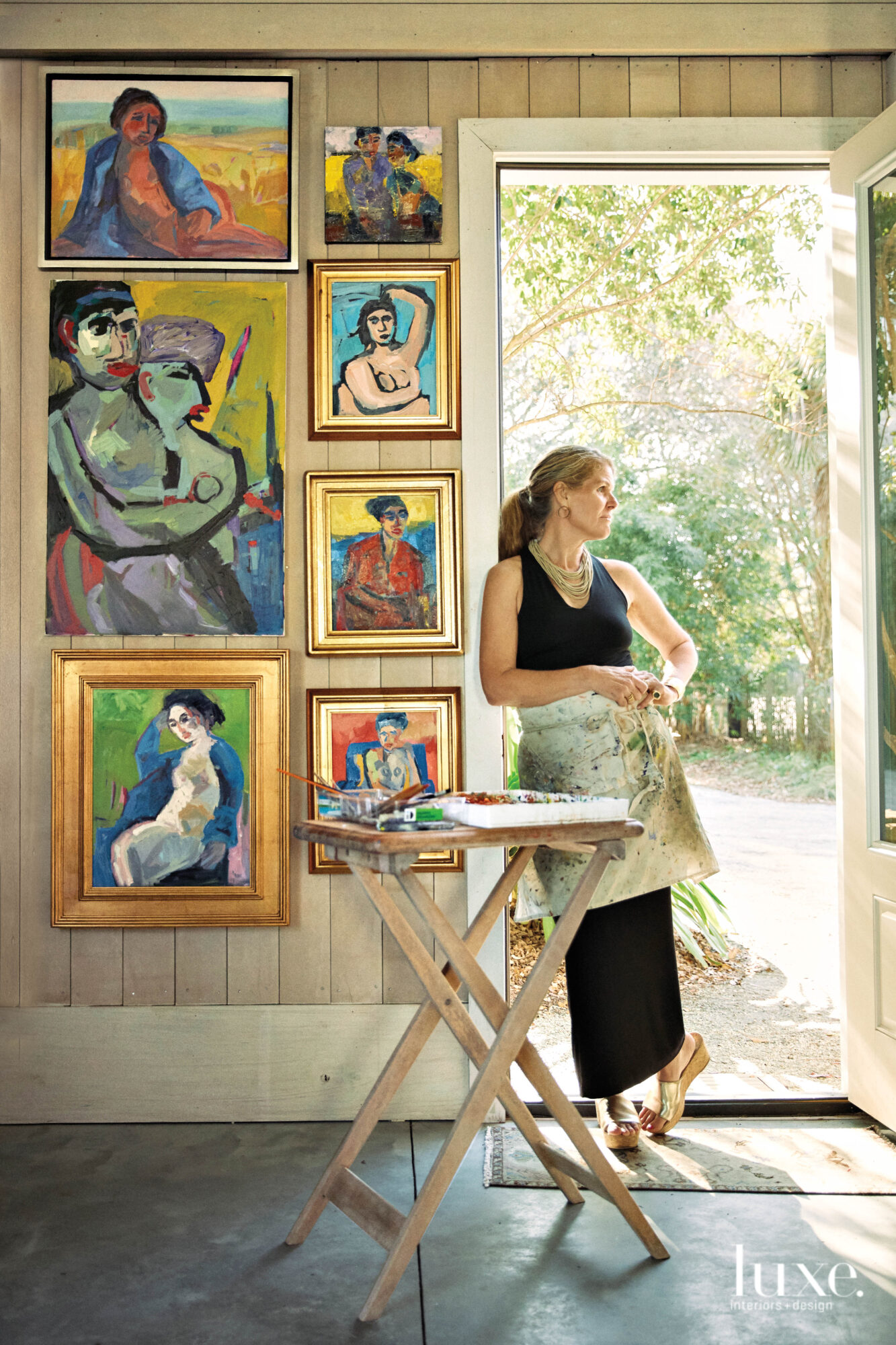 Woman wearing half apron, leaning against door frame, paintings on wall and folding table in foreground