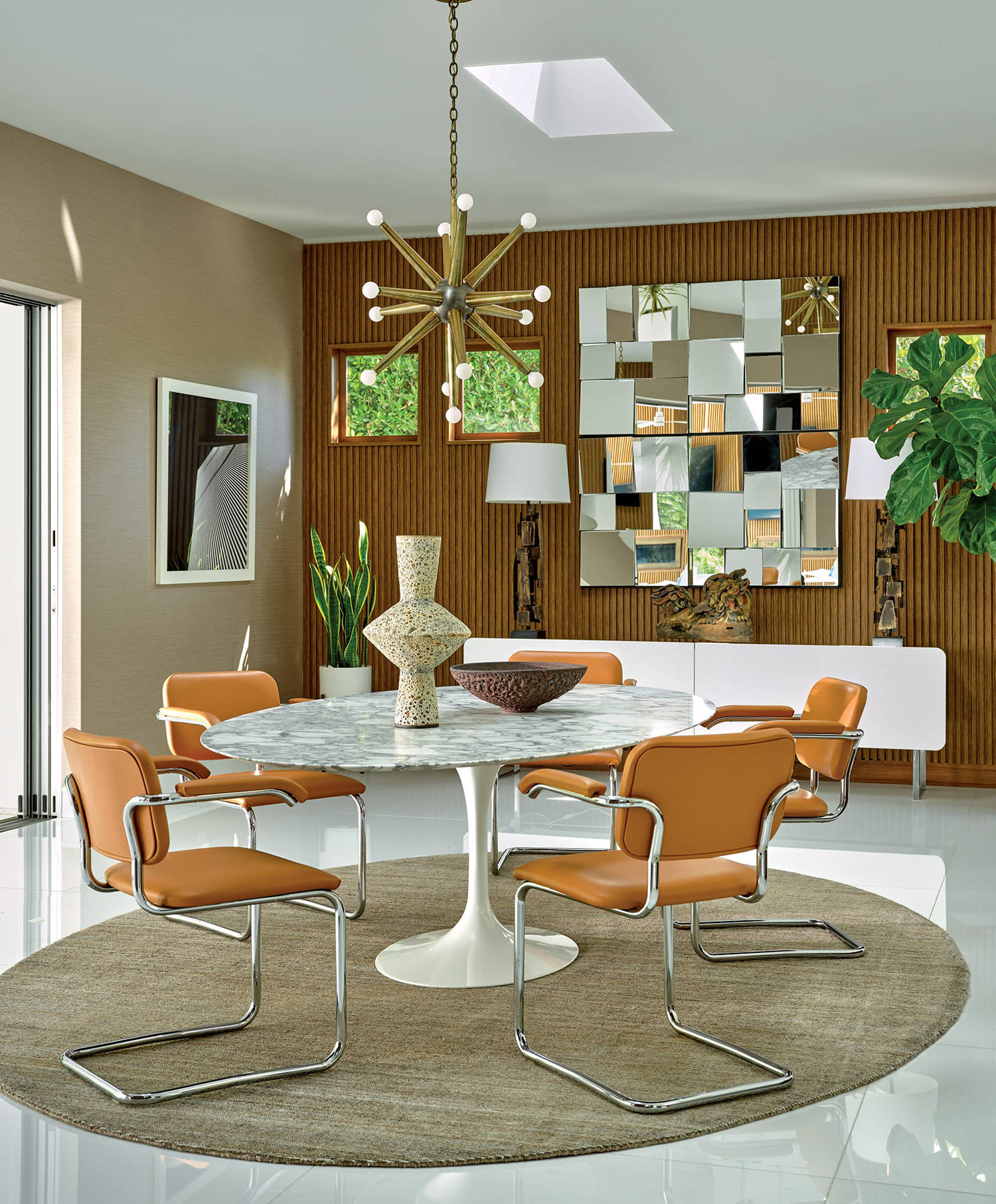 A white dining table surrounded by five chairs, in a room with a large mirror mounted on the wall and a chandelier hanging above.