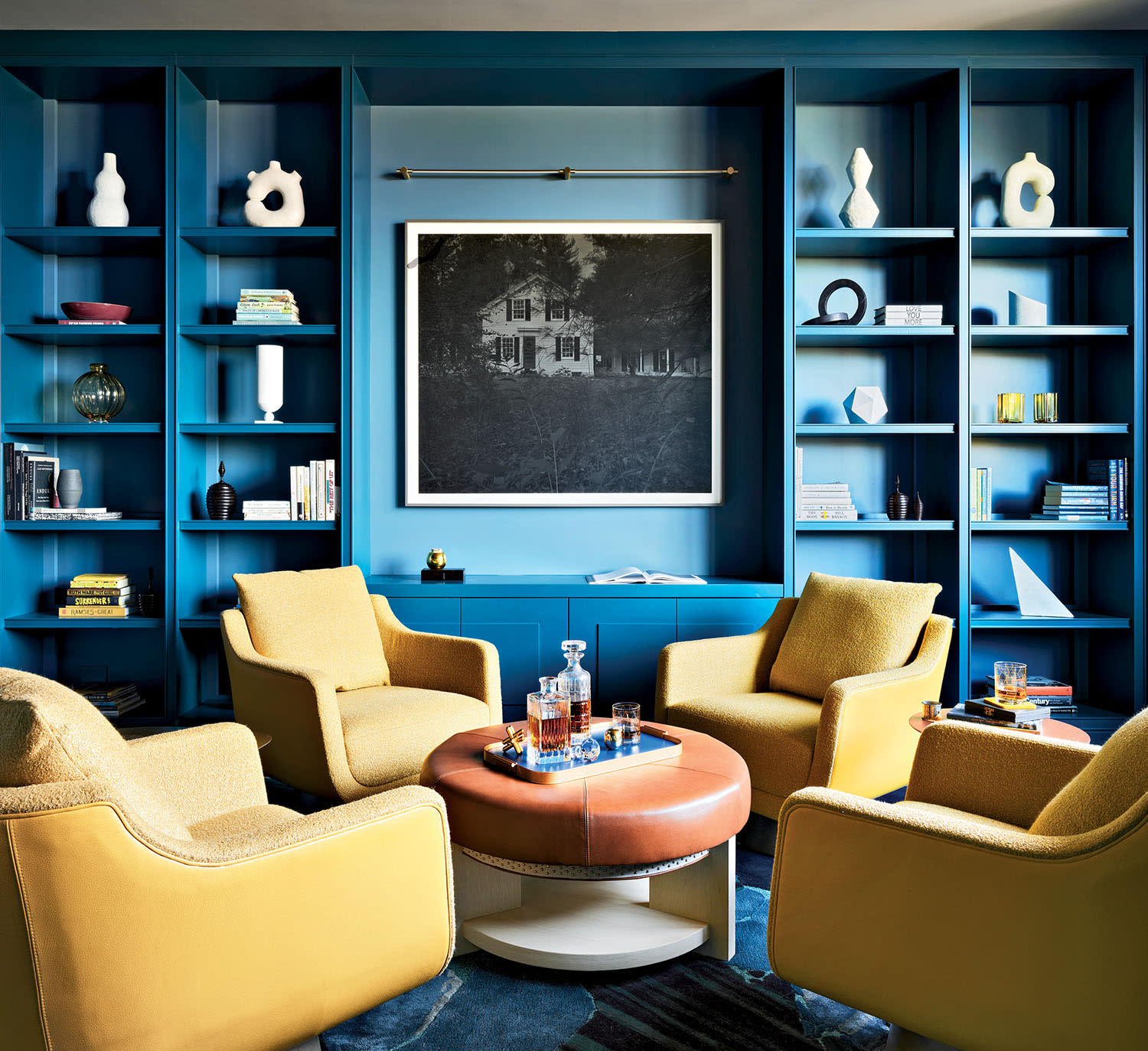 Cozy room with bright blue bookshelves framing a black-and-white photo. Four mustard yellow armchairs encircle a round brown ottoman, creating an inviting, stylish atmosphere.