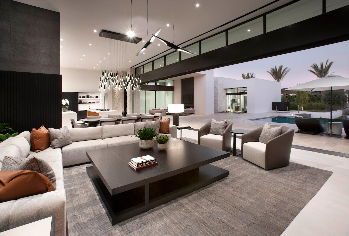 Modern living room with sleek furniture and neutral tones. A large sectional sofa surrounds a dark coffee table, and a glass door reveals a poolside view.