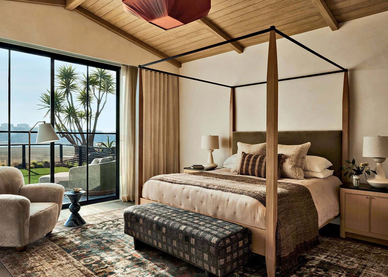 Stylish bedroom with a four-poster bed, plush chairs, and modern decor. Large window illuminates the space, offering a view of a sunny garden outside.