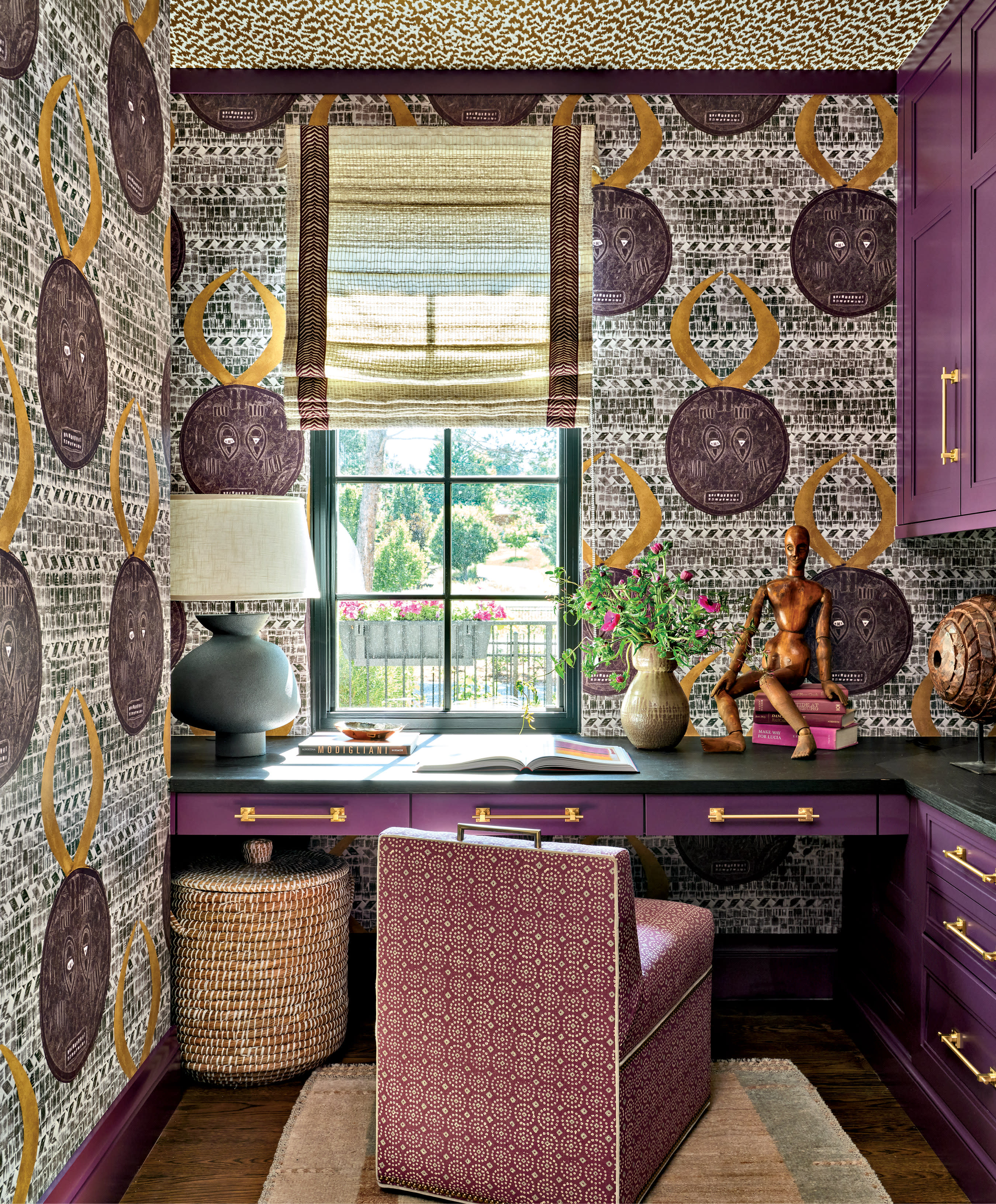 A vibrant home office with patterned wallpaper and a purple theme. The desk is adorned with books, a sculpture, and a vase of flowers. A large window offers a garden view, creating a lively and artistic atmosphere.
