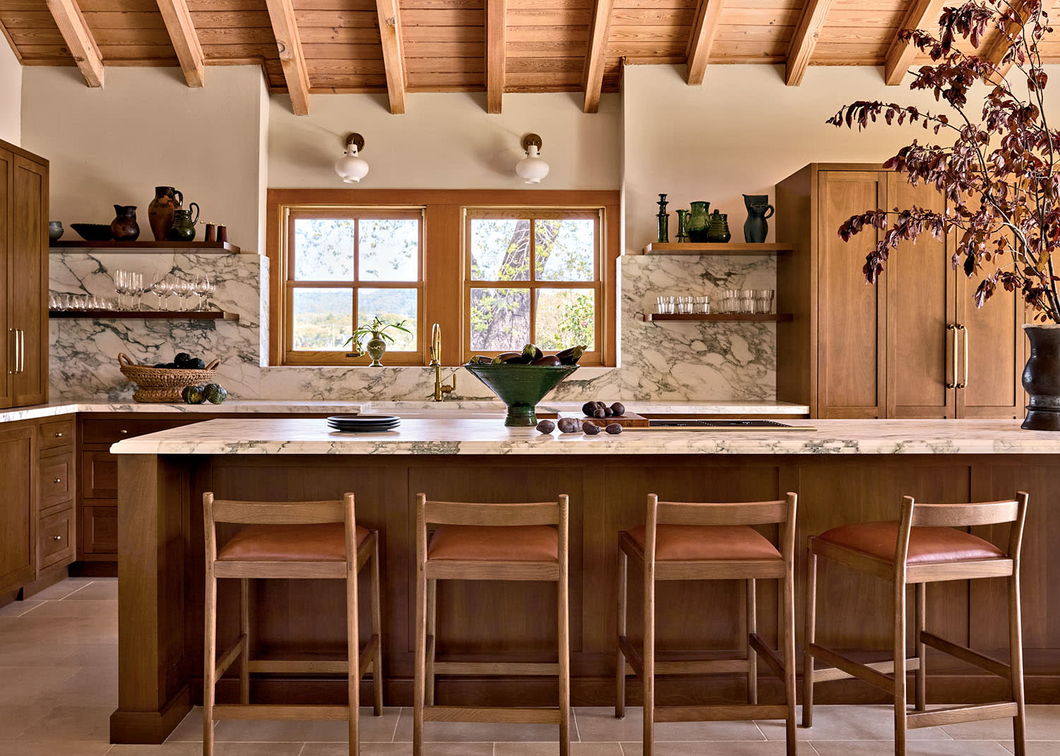 Warm, rustic kitchen with a wooden ceiling, marble countertops, and a central island with four chairs. Natural light streams through a large window.