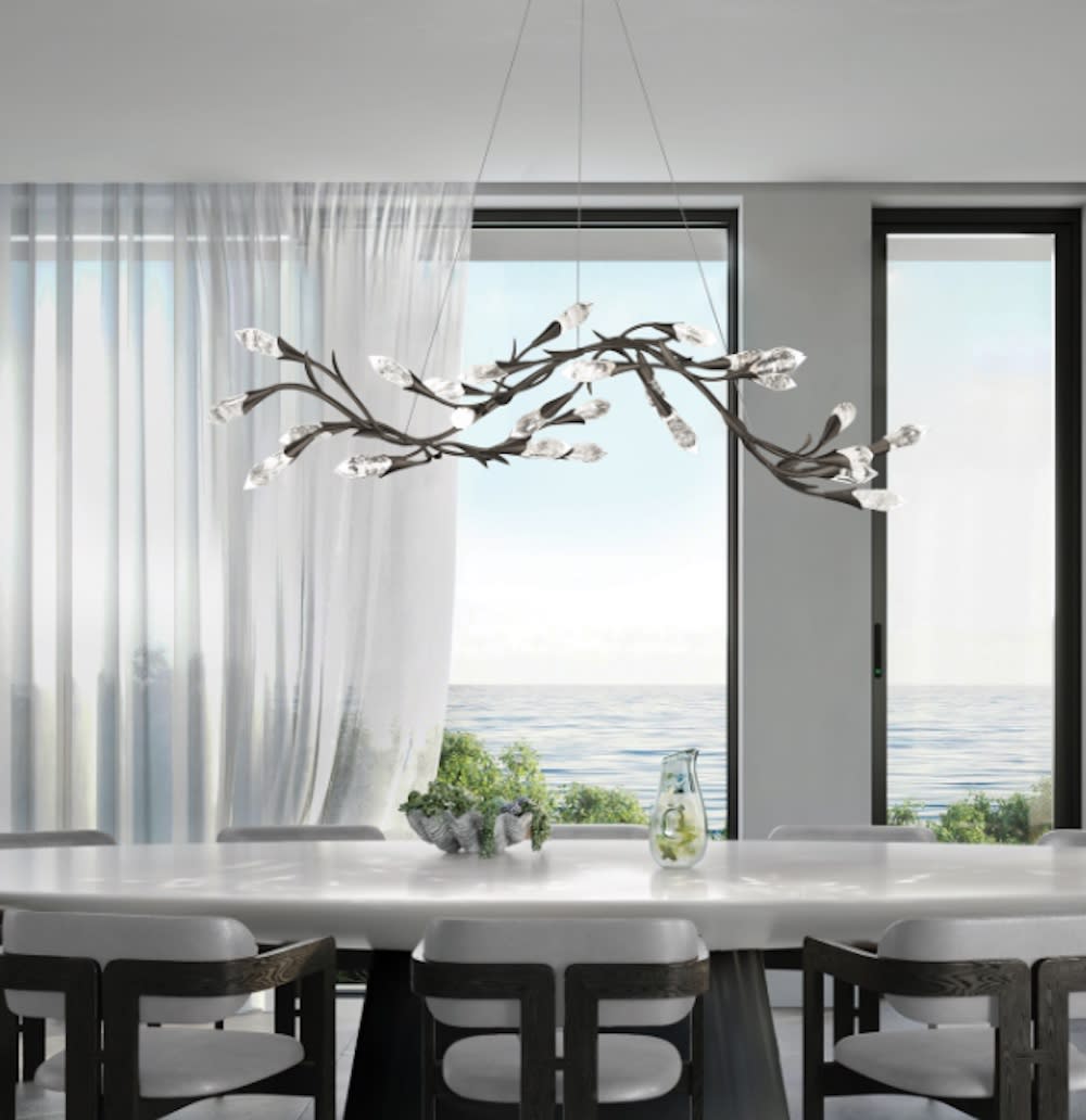 A contemporary dining room with a grand chandelier hanging above a round table, offering a scenic view of water through the window.