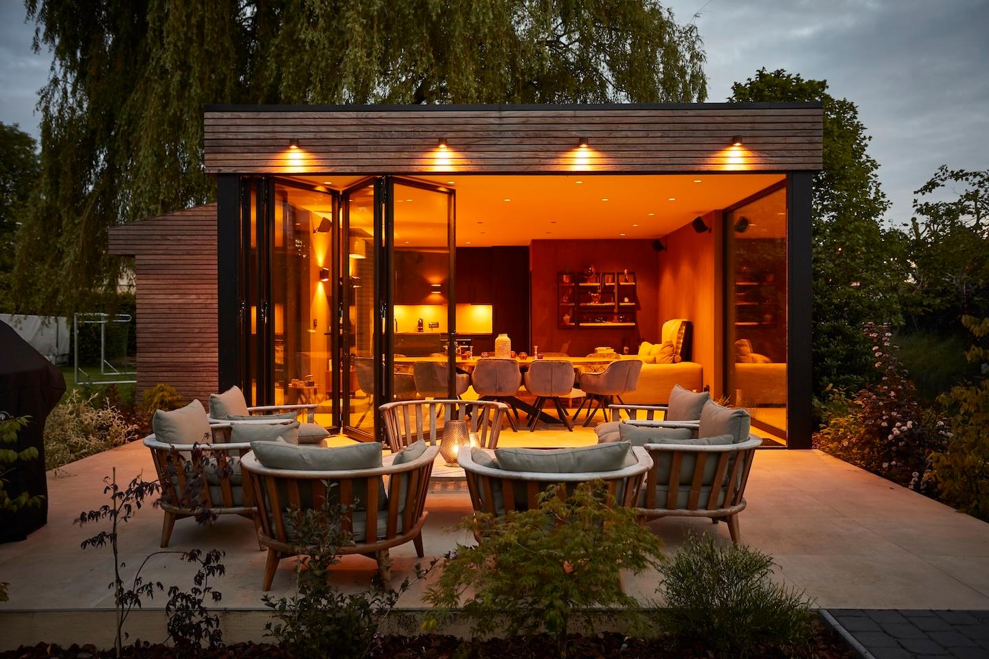 Modern patio with cozy chairs encircling a table, facing a warmly lit glass-walled interior. Evening ambiance, serene and inviting outdoor space.