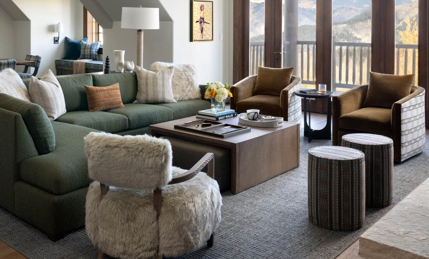 A cozy living room with a green L-shaped sofa, fluffy chair, and wooden coffee table adorned with books and flowers. Large windows offer a mountain view.