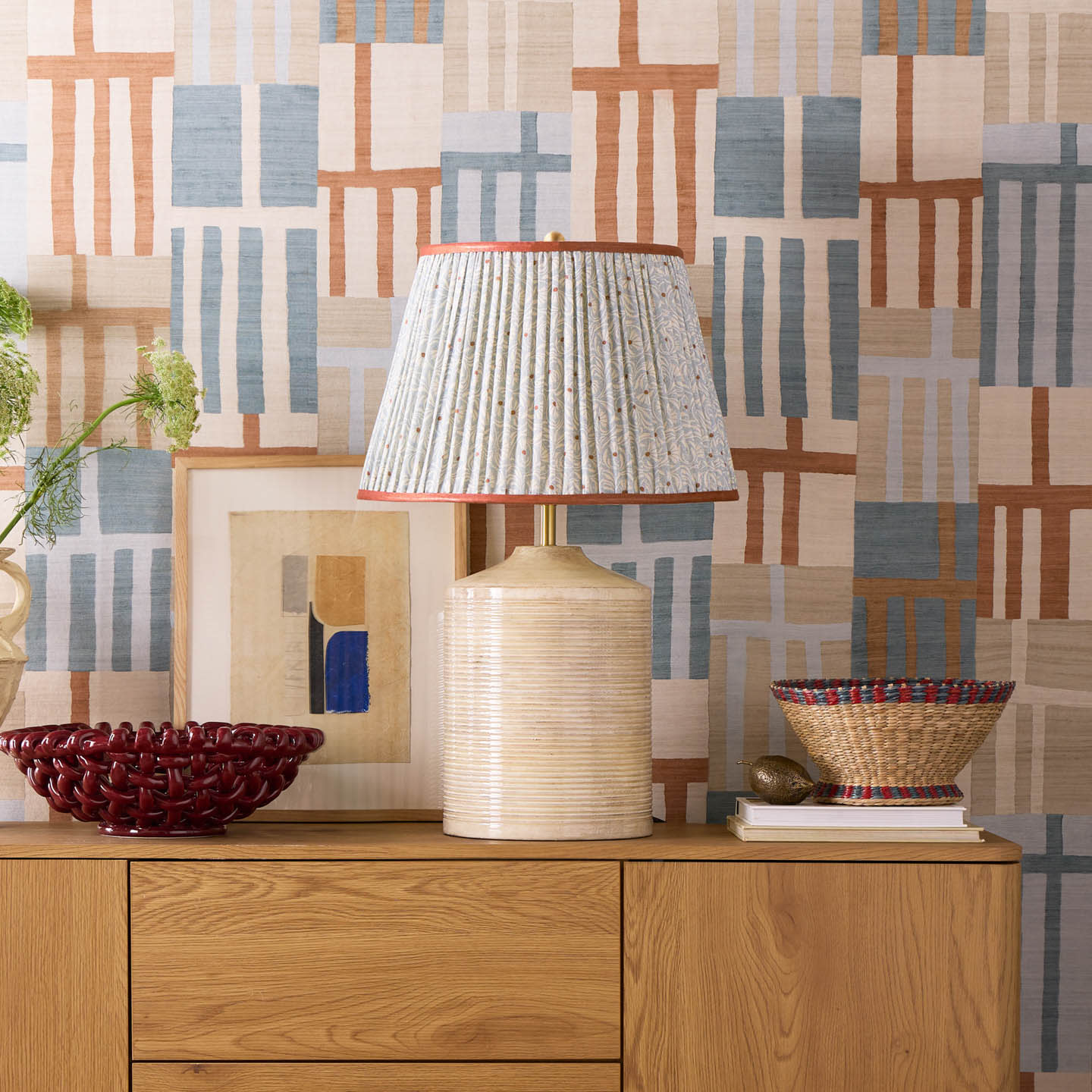 A lamp with a pleated beige shade sits on a wooden sideboard alongside a red bowl, artwork, and woven basket. 