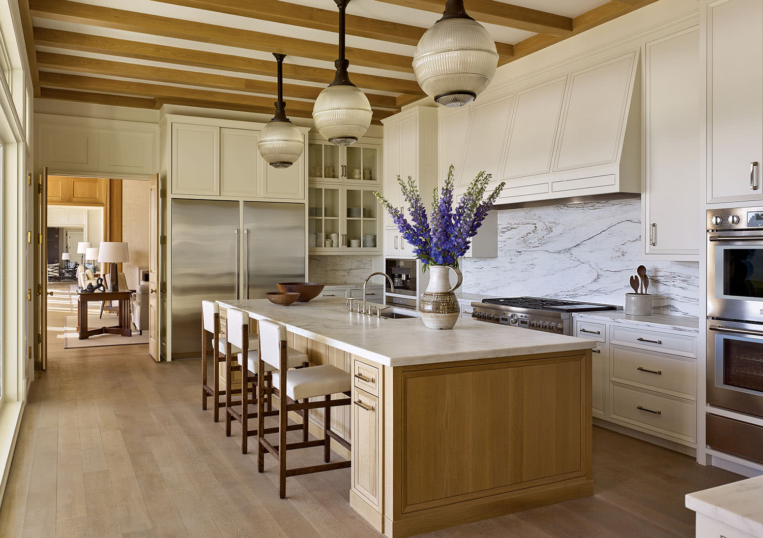 Elegant kitchen with white cabinetry, a large island with bar stools, and marble countertops. There are exposed wooden beams, pendant lights, and a vase of purple flowers.