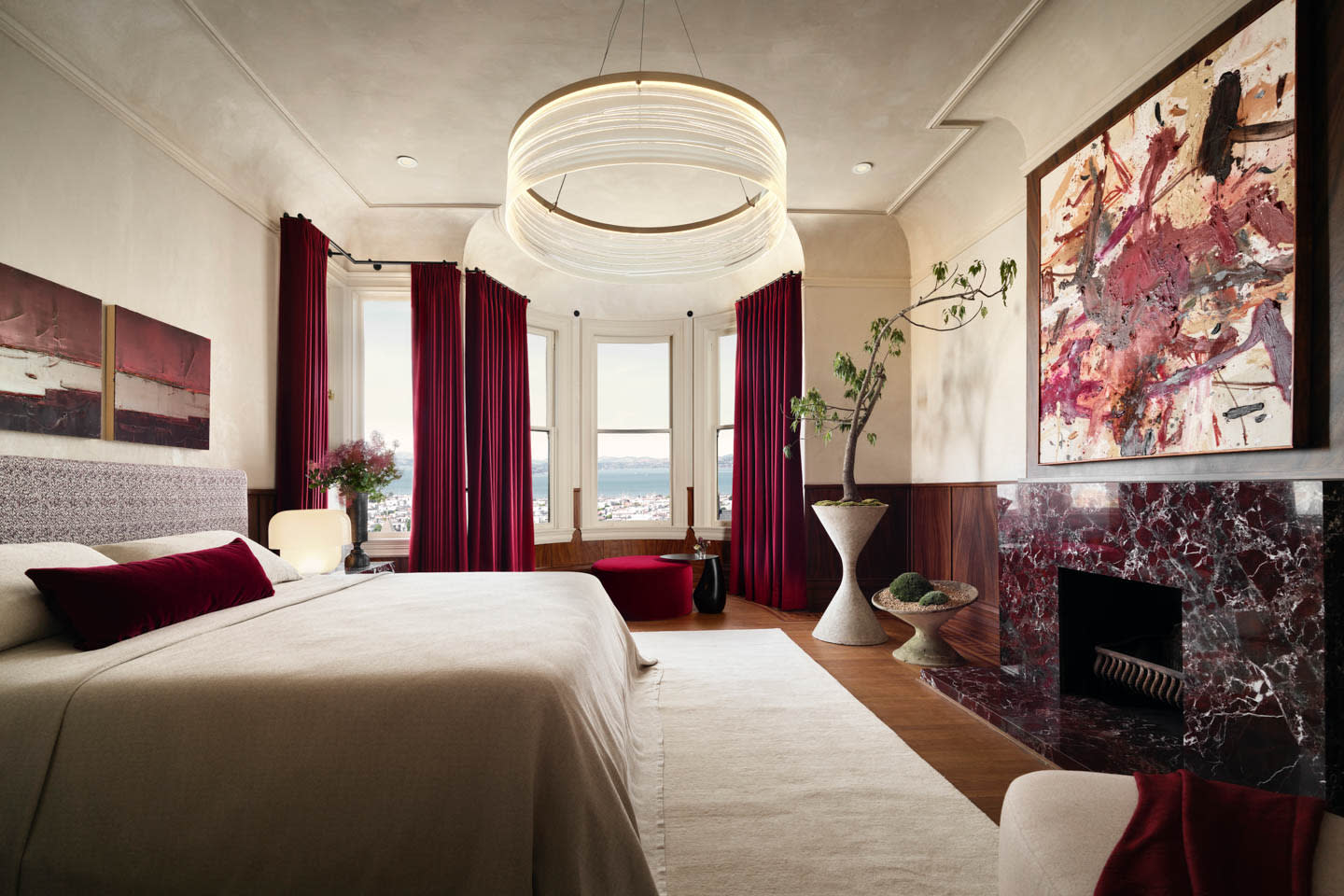Luxurious bedroom with large chandelier, abstract art, and rich maroon accents. Features a fireplace, plush bedding, and bay windows overlooking a scenic view.