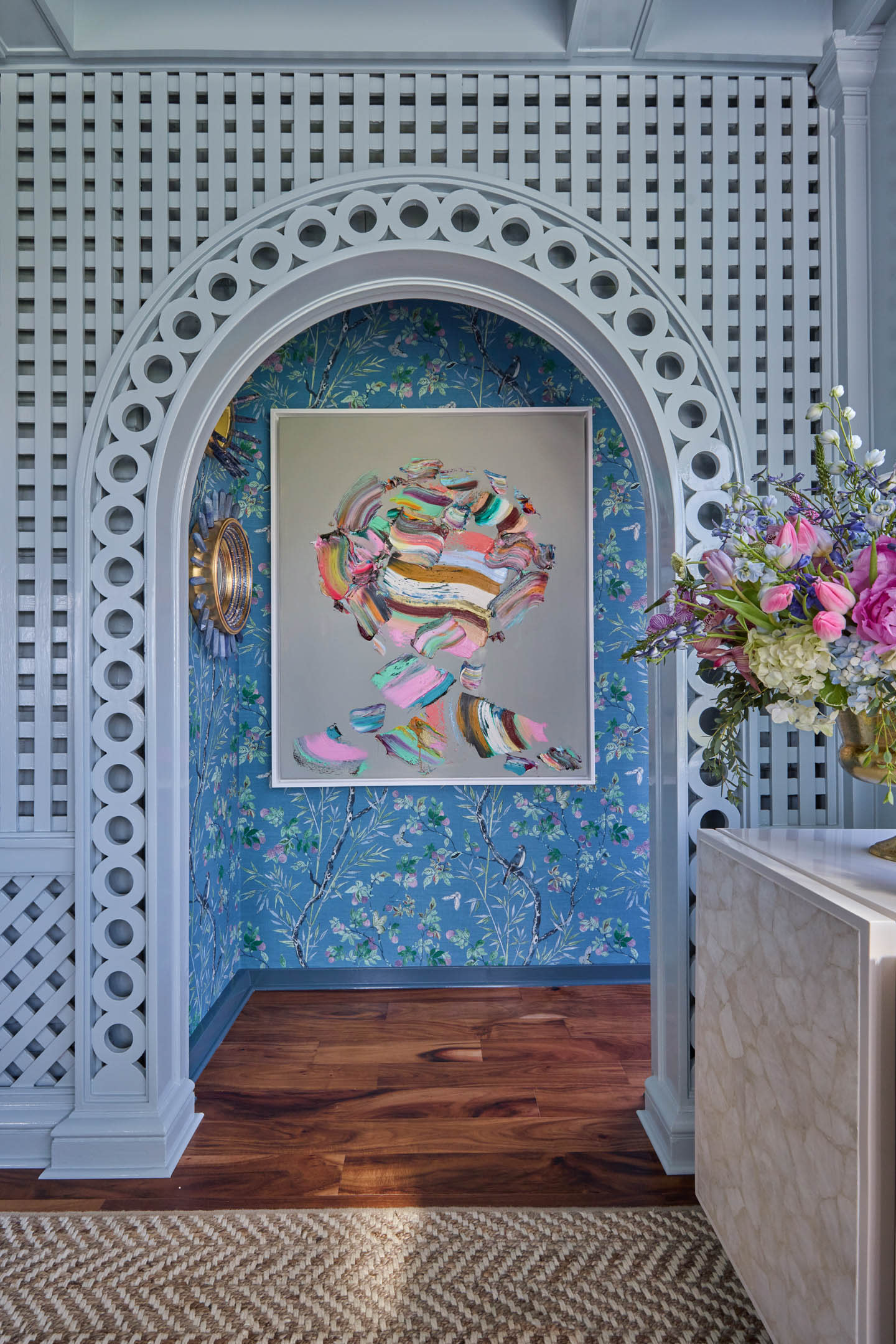 An ornate hallway features a colorful abstract painting set against floral blue wallpaper. The mood is vibrant and elegant, complemented by fresh flowers.