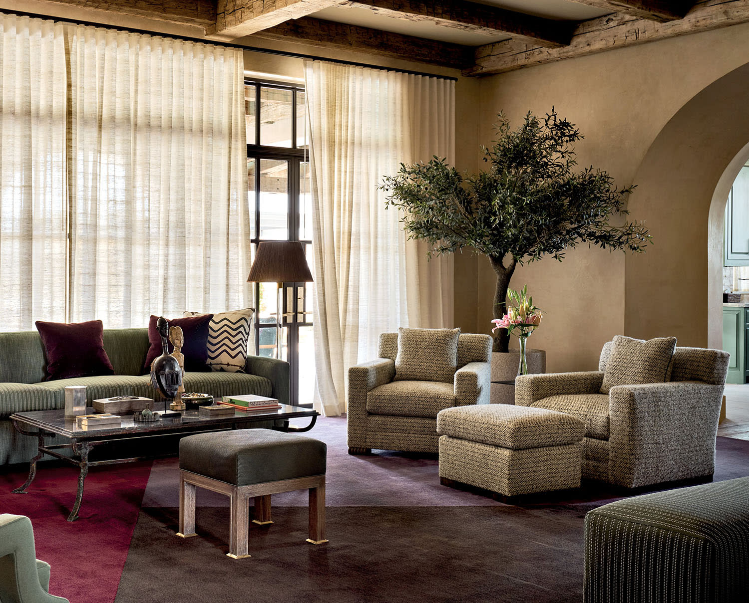 Elegant living room with textured sofas and chairs, rich purple rug, and large potted plant. Sunlight filters through sheer curtains, casting a warm glow. Rustic wood beams add a cozy charm.
