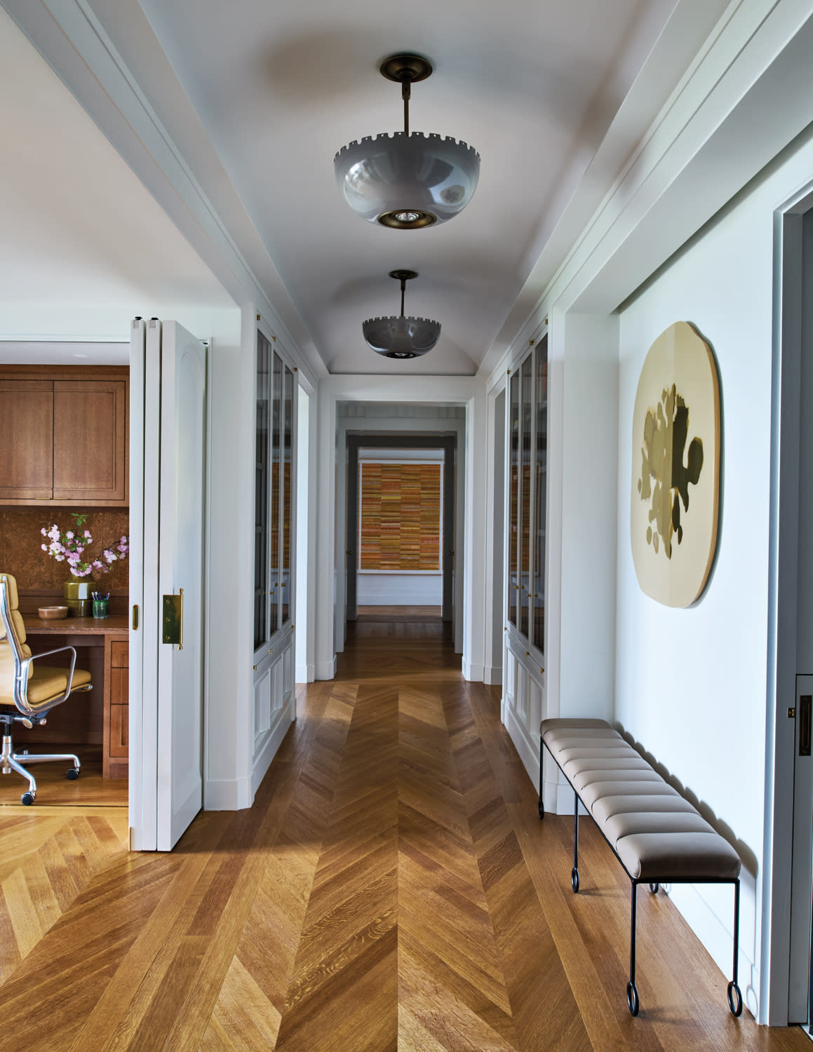 A hallway with herringbone wood flooring, modern pendant lights, and a wall artwork. On the right, a tufted bench sits below a minimalist painting.