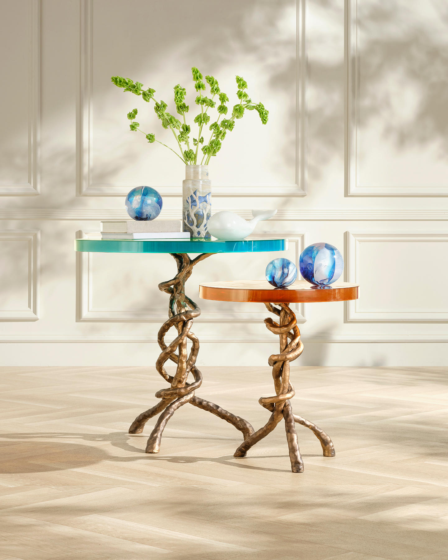 Two ornate tables with twisted bronze bases and colorful tops are adorned with glass spheres, a vase of green flowers, and a white teapot. Sunlight casts soft shadows, evoking a serene atmosphere.