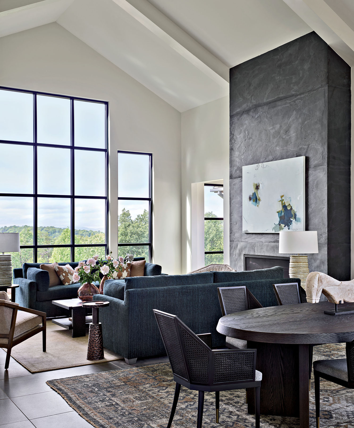 A living room with sofas next to tall windows with mountain views. A dining area sits nearby.