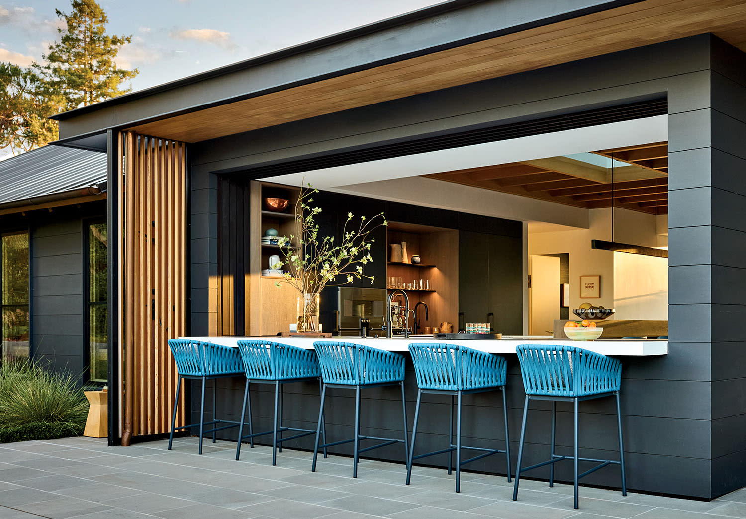 Modern outdoor kitchen with a sleek black exterior, large open window, and blue chairs at a counter. Warm interior lighting creates a cozy ambiance.
