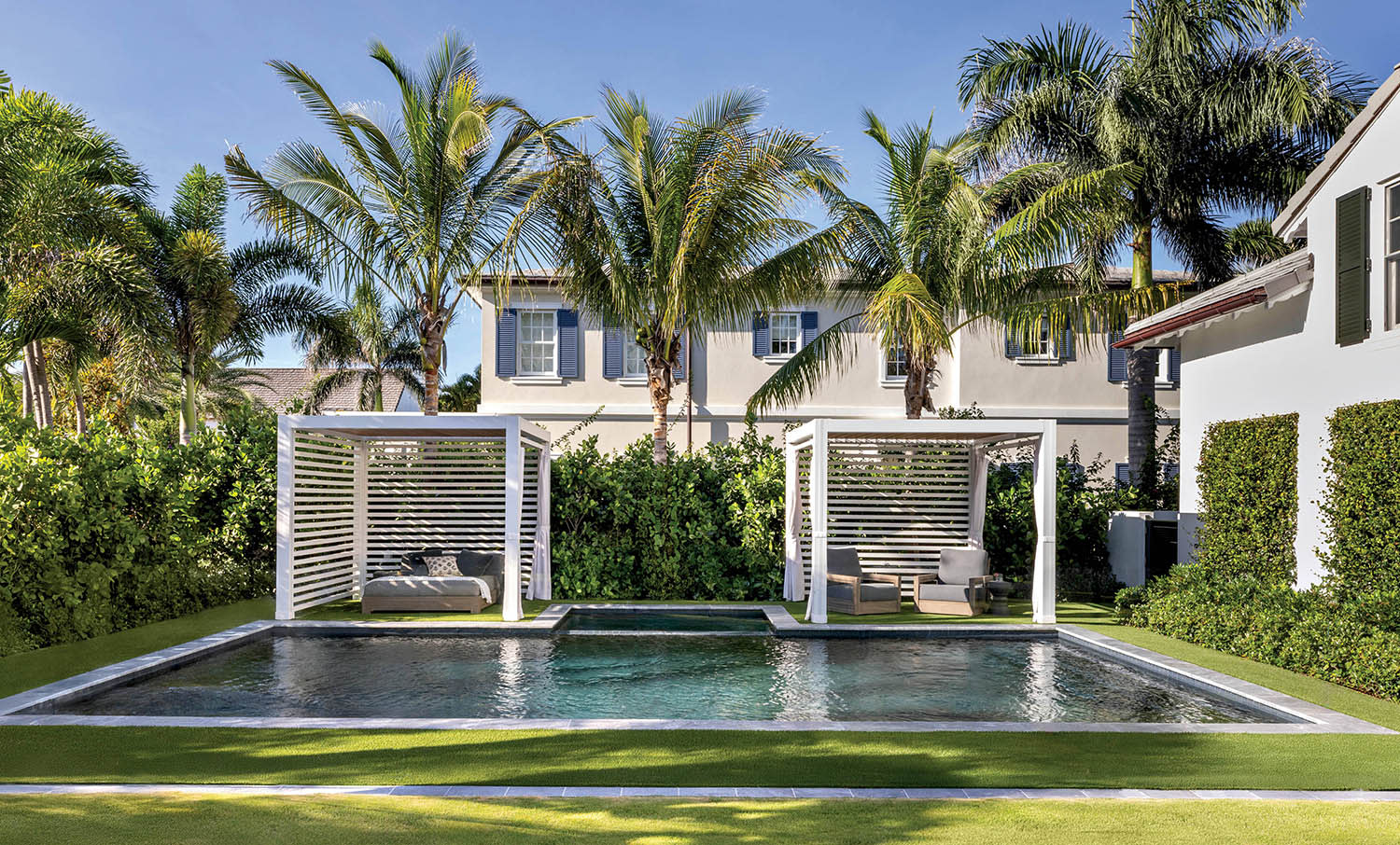 In-ground pool flanked by two white cabanas with seating surrounded by a lawn and palm trees.