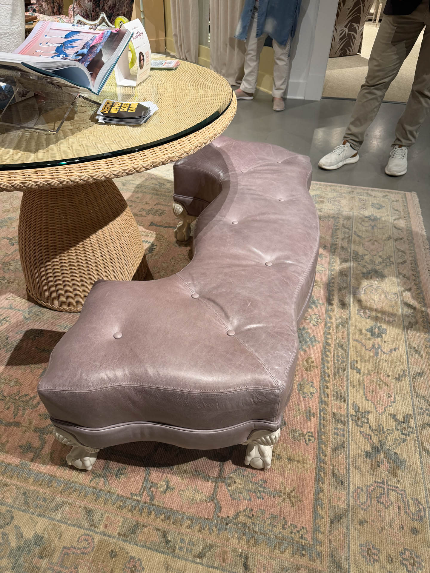 A curved, tufted pink leather bench with ornate white legs is positioned near a round wicker table displaying magazines. The setting is on a patterned rug.