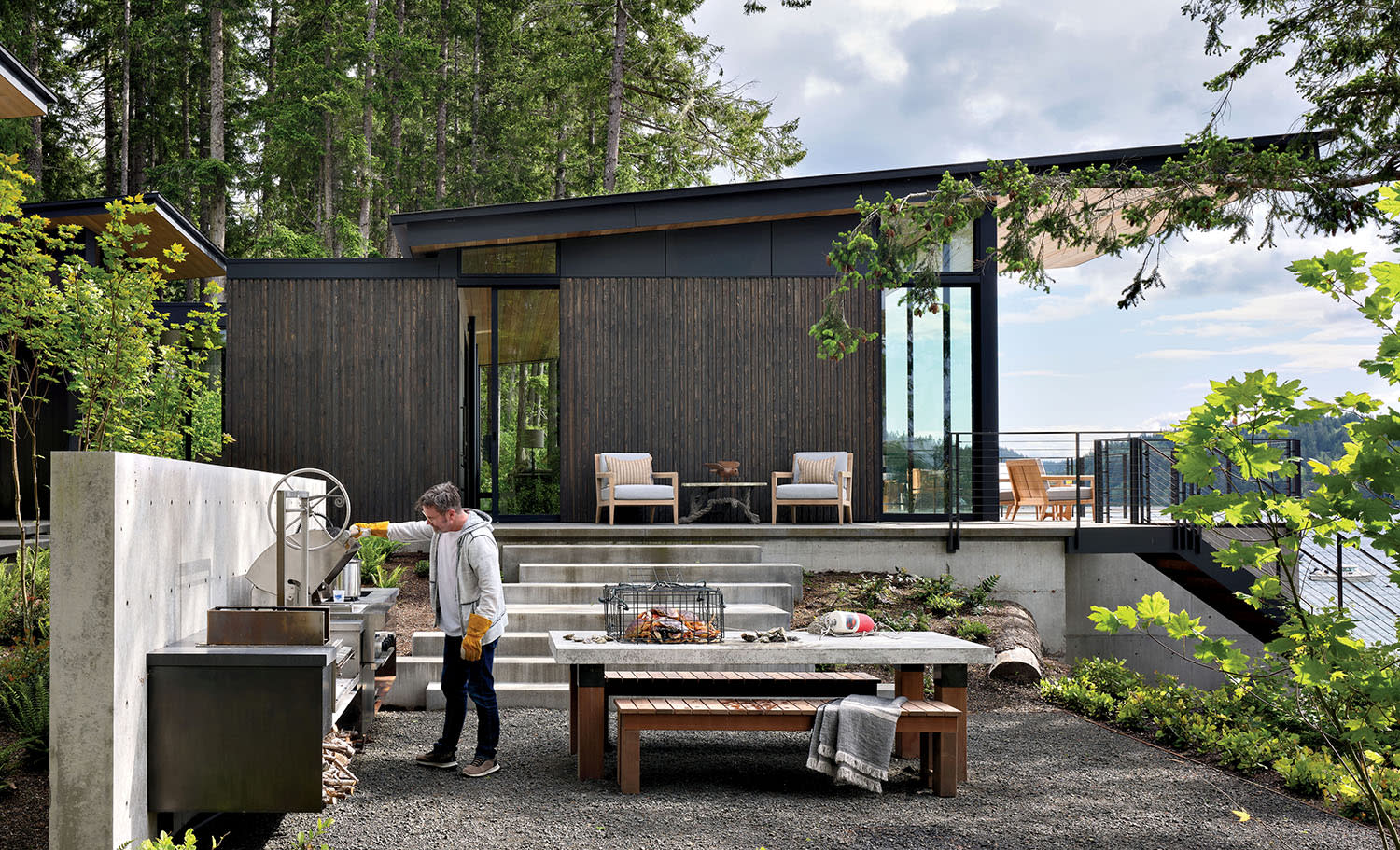 Modern wood-clad home with large windows, surrounded by trees. A person uses an outdoor kitchen with a grill and picnic table. The scene feels serene and nature-focused.