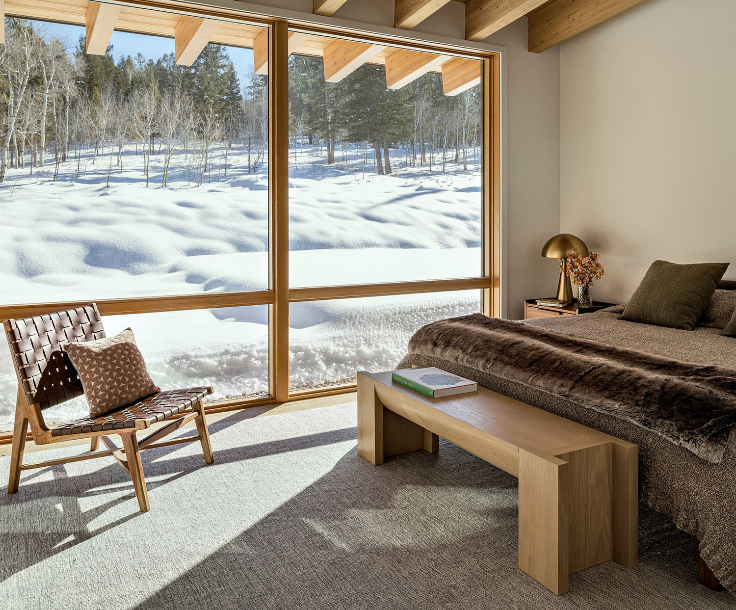 A bed with a wood bench at the foot and a nearby armchair set near large windows looking out at snow.
