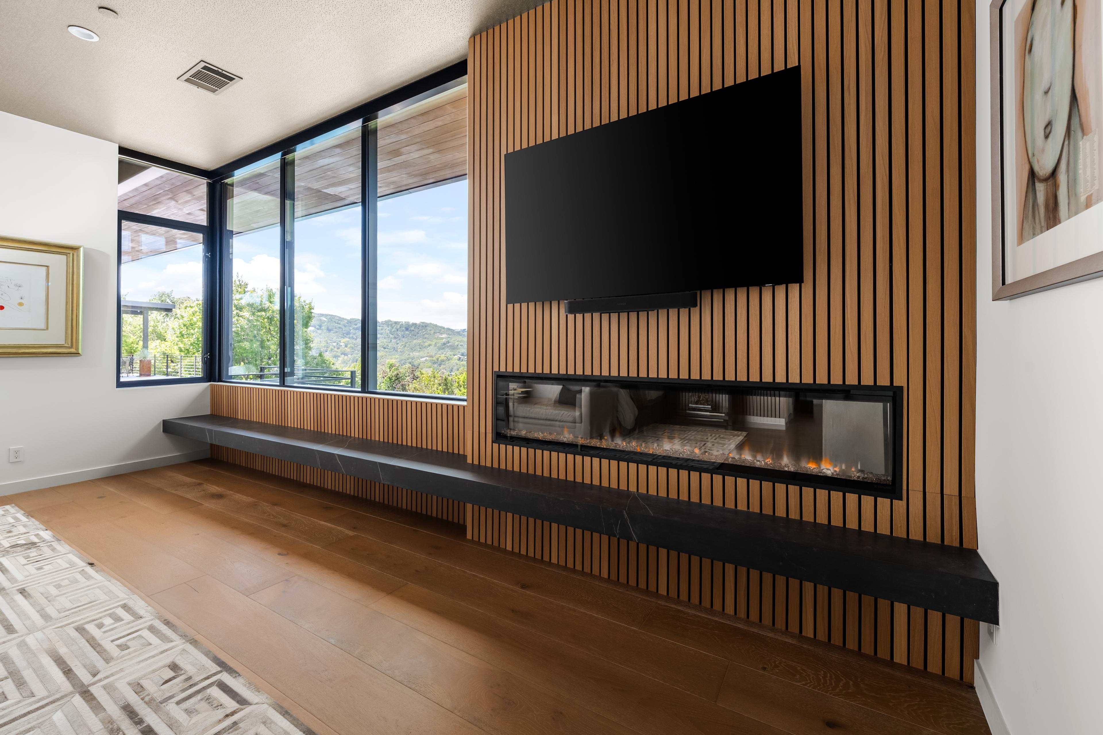 This modern living space features a sleek slatted wood accent wall with a linear gas fireplace and mounted TV. Expansive windows invite natural light and stunning views, while warm wood floors complete the contemporary design.