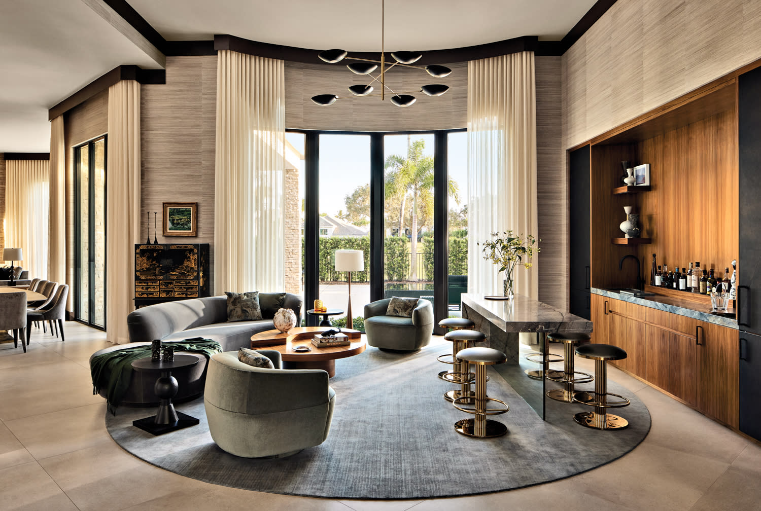Luxurious living room with modern decor features a circular rug, plush seating, a sleek bar, and large windows. Soft light and muted tones convey elegance.