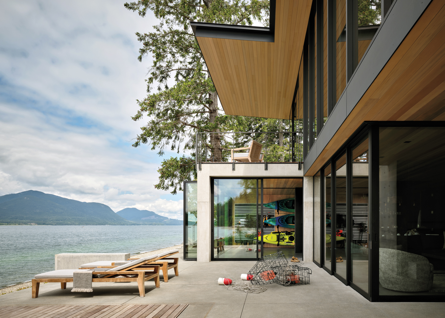 Modern lakeside house with large glass windows, wooden decks, and kayaks visible inside. Overcast sky adds a tranquil, serene atmosphere.