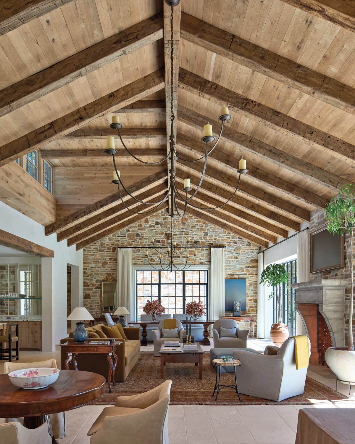Spacious living room with vaulted wood-beamed ceiling, rustic stone wall, and large window. Cozy seating, elegant chandeliers, and a warm, inviting atmosphere.