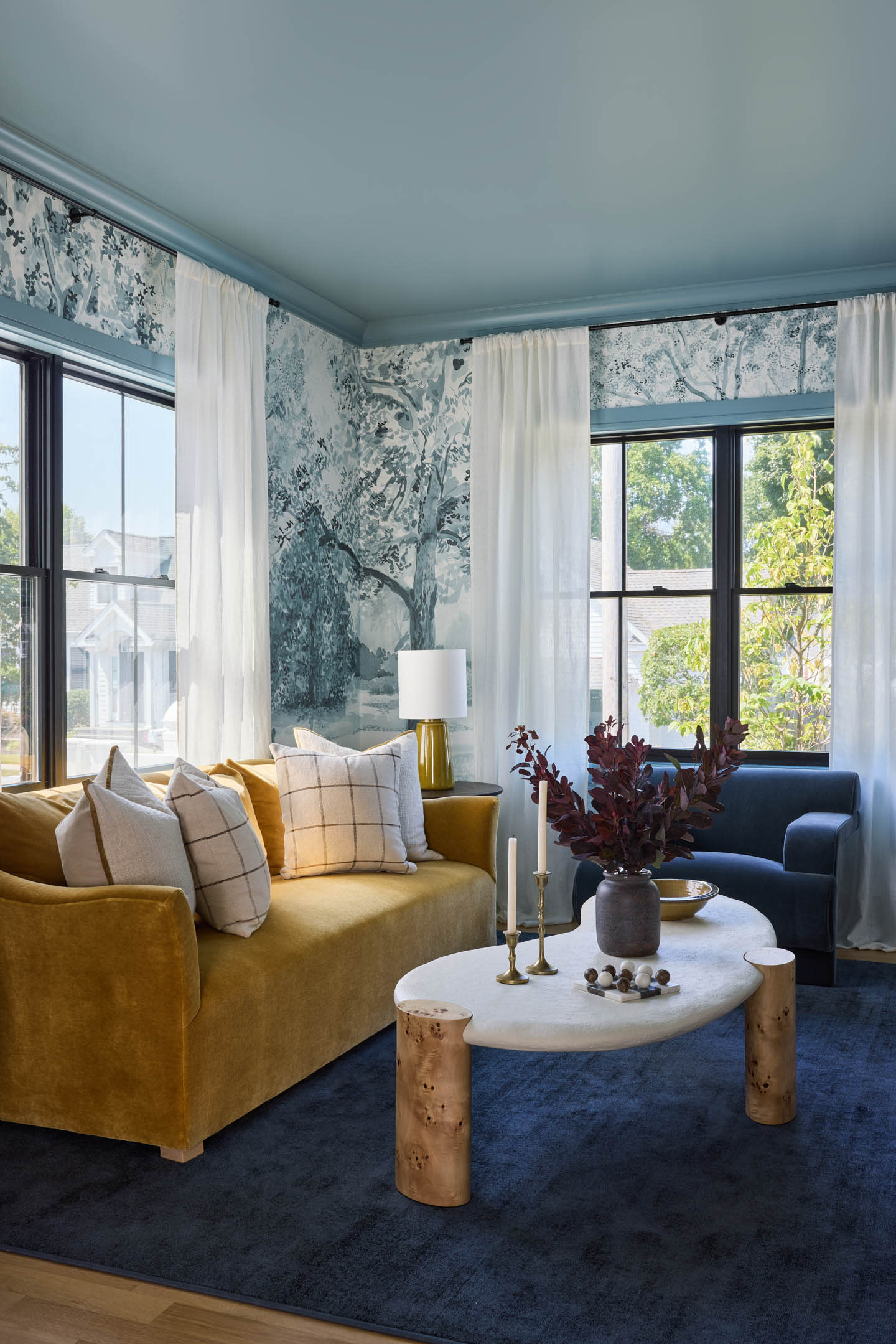 A cozy living room features a mustard-yellow sofa with plaid pillows, a blue armchair, a marble coffee table with candles, and leafy wallpaper accents.