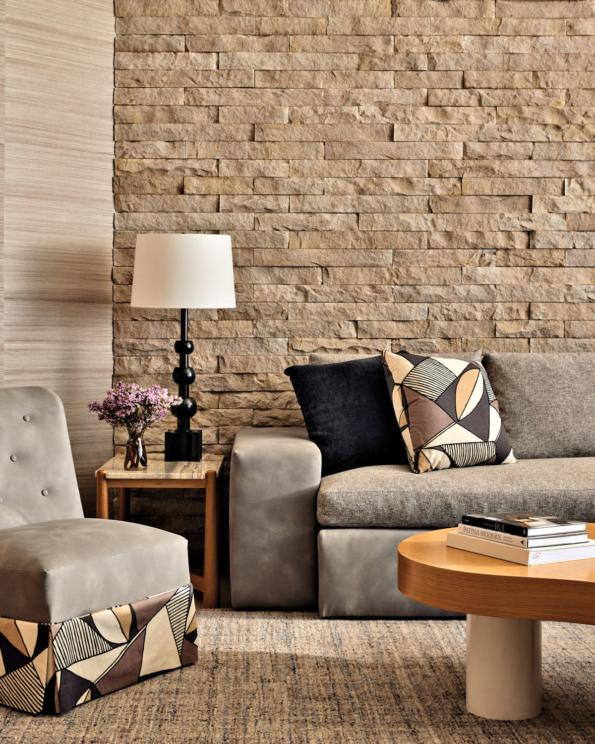 Cozy living room with a stone accent wall, gray sofa, geometric pillows, side chair, wooden coffee table, modern lamp, and vase with purple flowers.