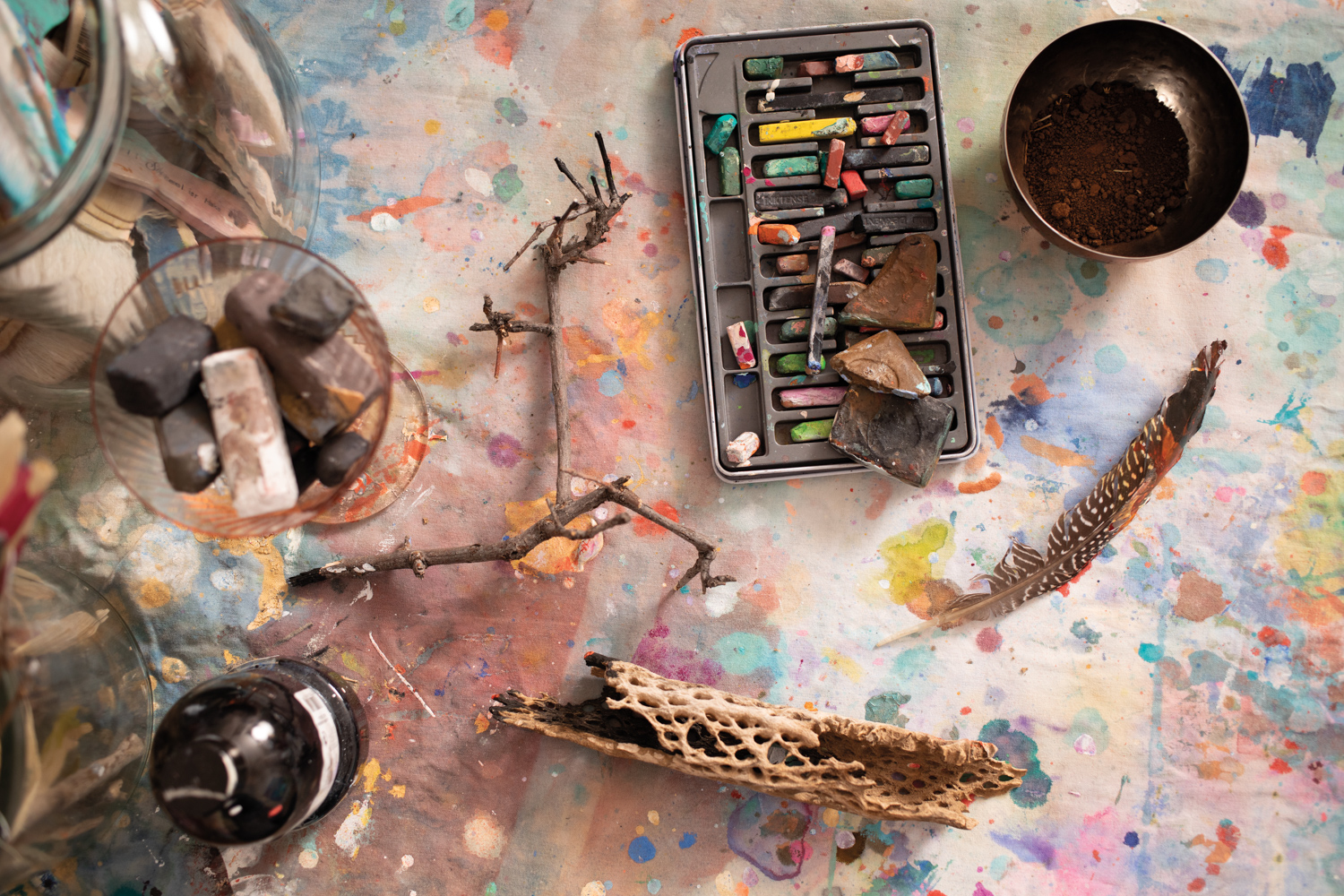 pastels, paint, a bowl of dirt, driftwood and a feather sit on a paint-splattered table