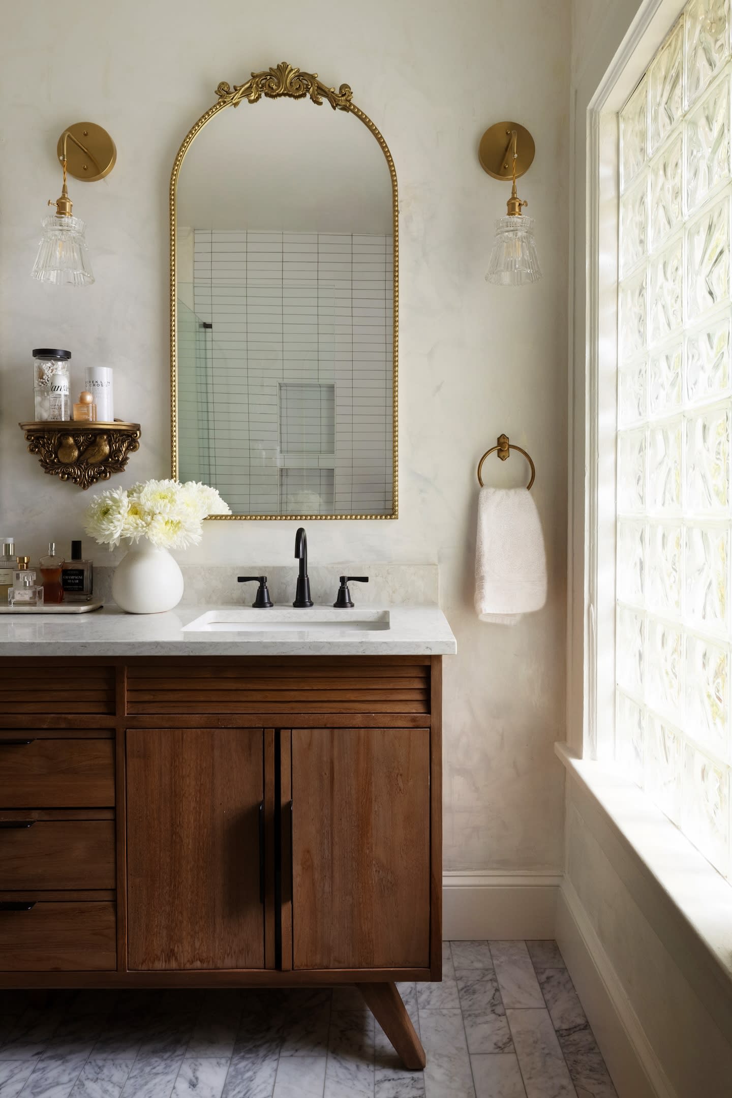 Elegant bathroom with a wooden vanity, marble countertop, and gold-framed mirror. Glass sconces, vase of flowers, and a towel on the right evoke a cozy, stylish atmosphere.