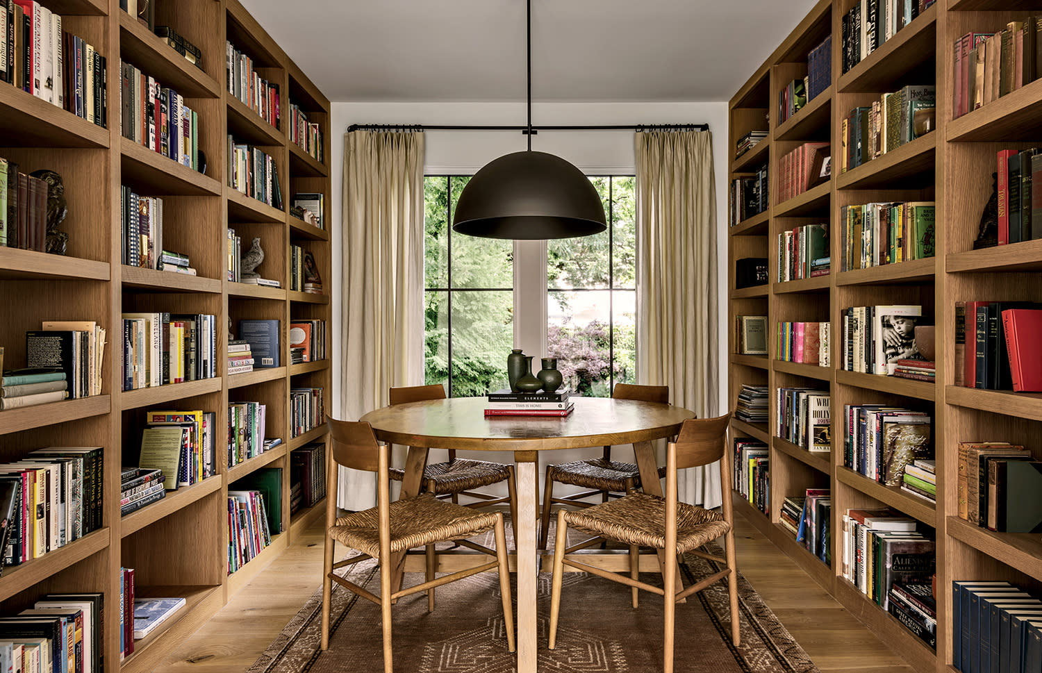 A cozy library with wooden bookshelves full of books lines both sides. A round table with chairs sits under a large pendant lamp next to a window with curtains.