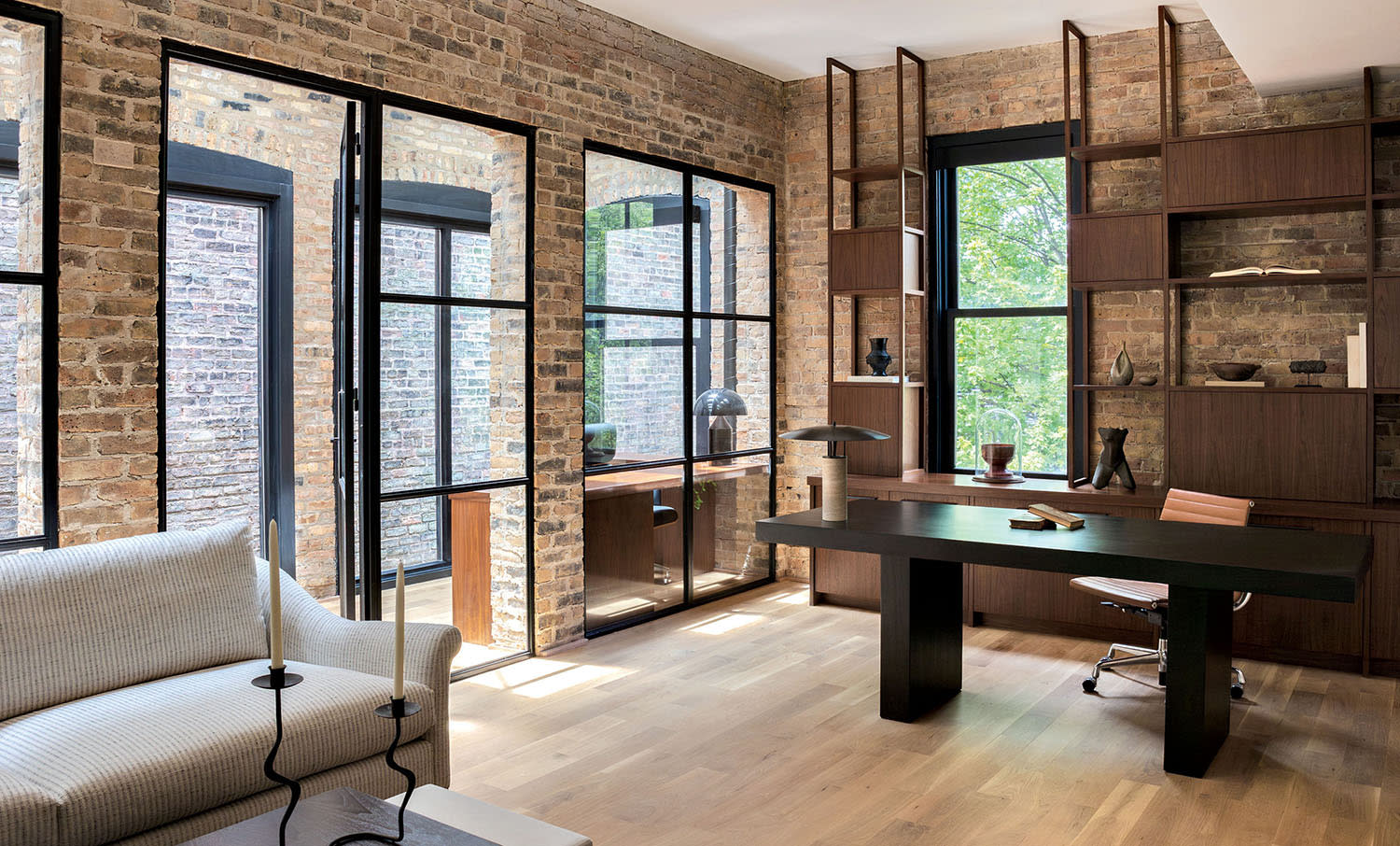A brick office area with hardwood floors, a gray couch in a corner and a desk with shelves behind it.